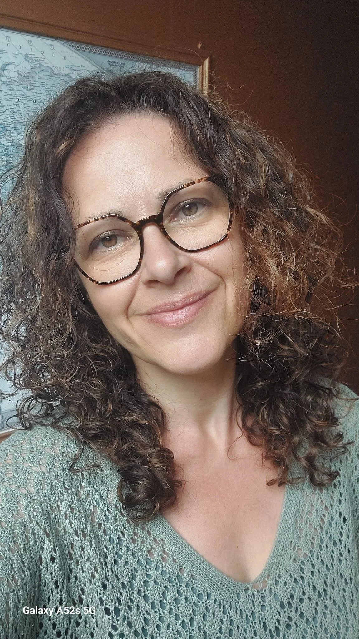 Une femme aux cheveux bouclés et bruns, portant des lunettes rondes et un pull en crochet vert, sourit devant un mur avec une carte du monde.