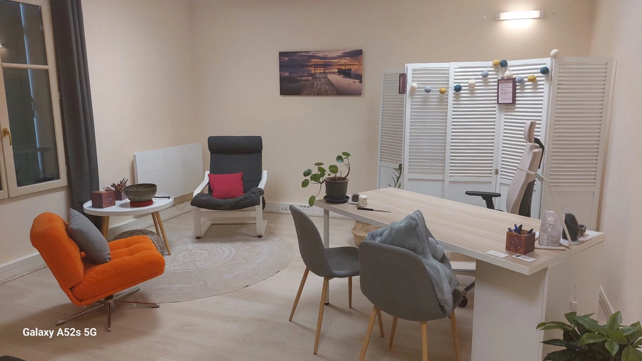 Une pièce de salon avec plusieurs fauteuils, une table de réunion, une étagère de séparation, un tableau représentant un quai de port au coucher du soleil, des plantes et diverses décorations.