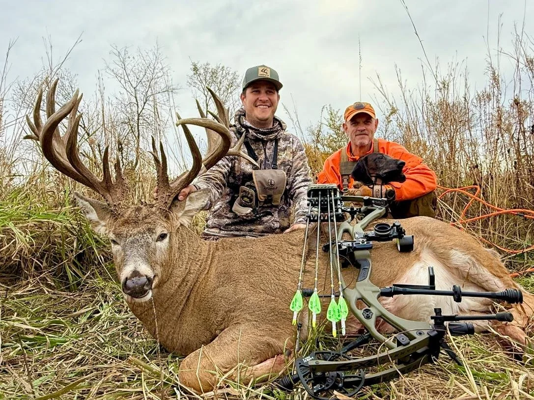 Two hunters, one with a camouflage jacket and another with an orange jacket, are posing in a field with a large deer with antlers. The deer is lying on the ground with a crossbow nearby. One of the hunters holds the deer's antlers, and a dog sits next to them. The background features tall grass and leafless trees.