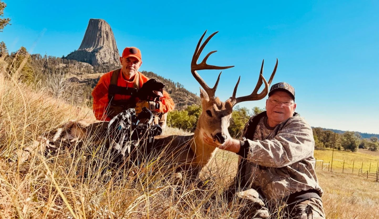 Two men on a grassy field with a large buck with antlers, mountains and blue sky in the background.