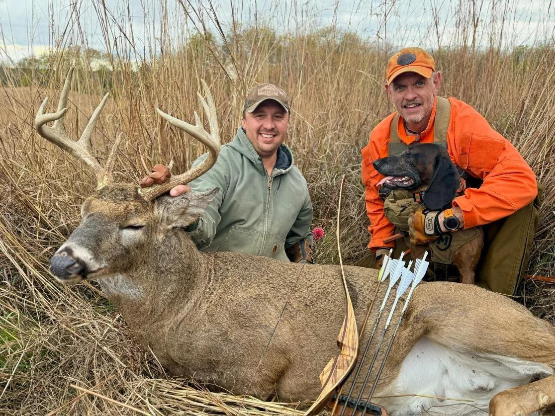Two men and a hunting dog pose with a killed deer in a field of tall grasses. One man holds the deer's antlers, and the other is kneeling beside the dog, all smiling at the camera.