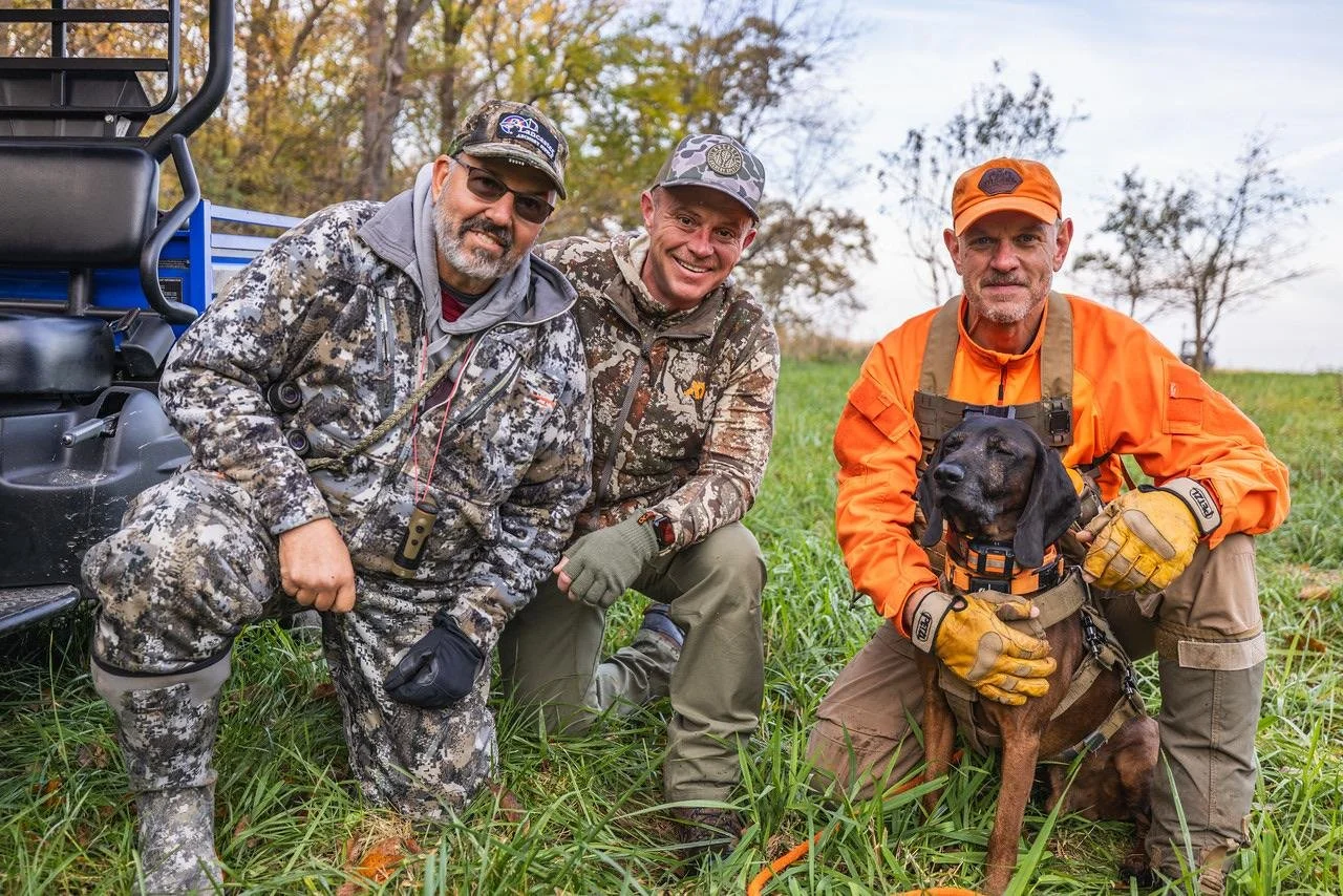 Three men in camouflage and orange hunting gear with a black hunting dog sitting on grass, trees in the background.
