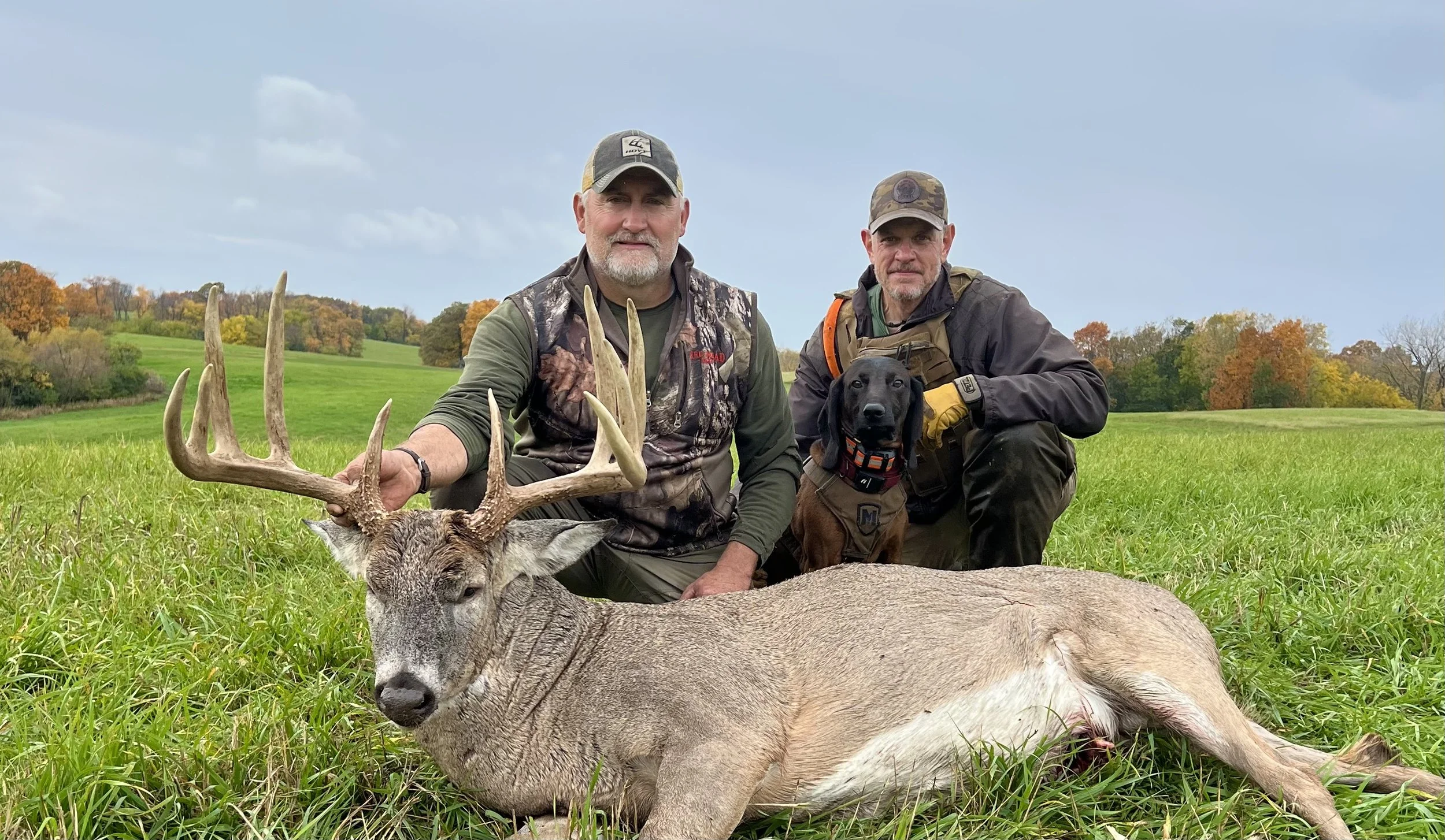 Two hunters and a hunting dog kneel beside a large dead deer with antlers in a grassy field, with trees displaying fall foliage in the background.