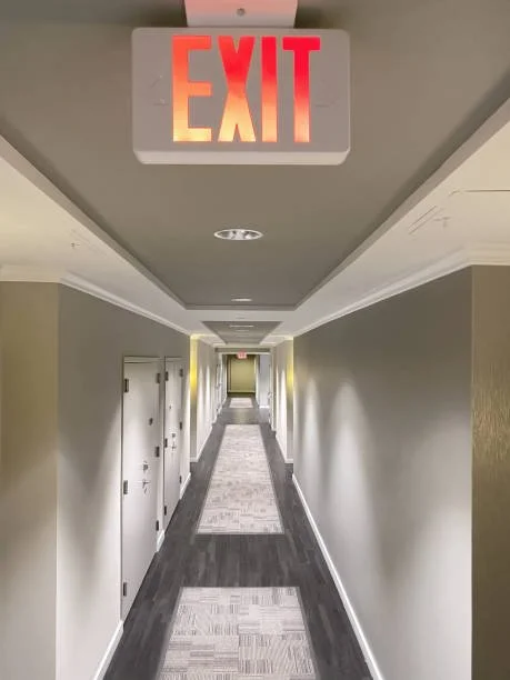 Long hotel corridor with multiple doors on each side, gray walls, and a patterned carpet. An illuminated red and orange 'EXIT' sign is mounted on the ceiling at the forefront. part of a Complete Fire and Security System in Southampton, Hampshire.