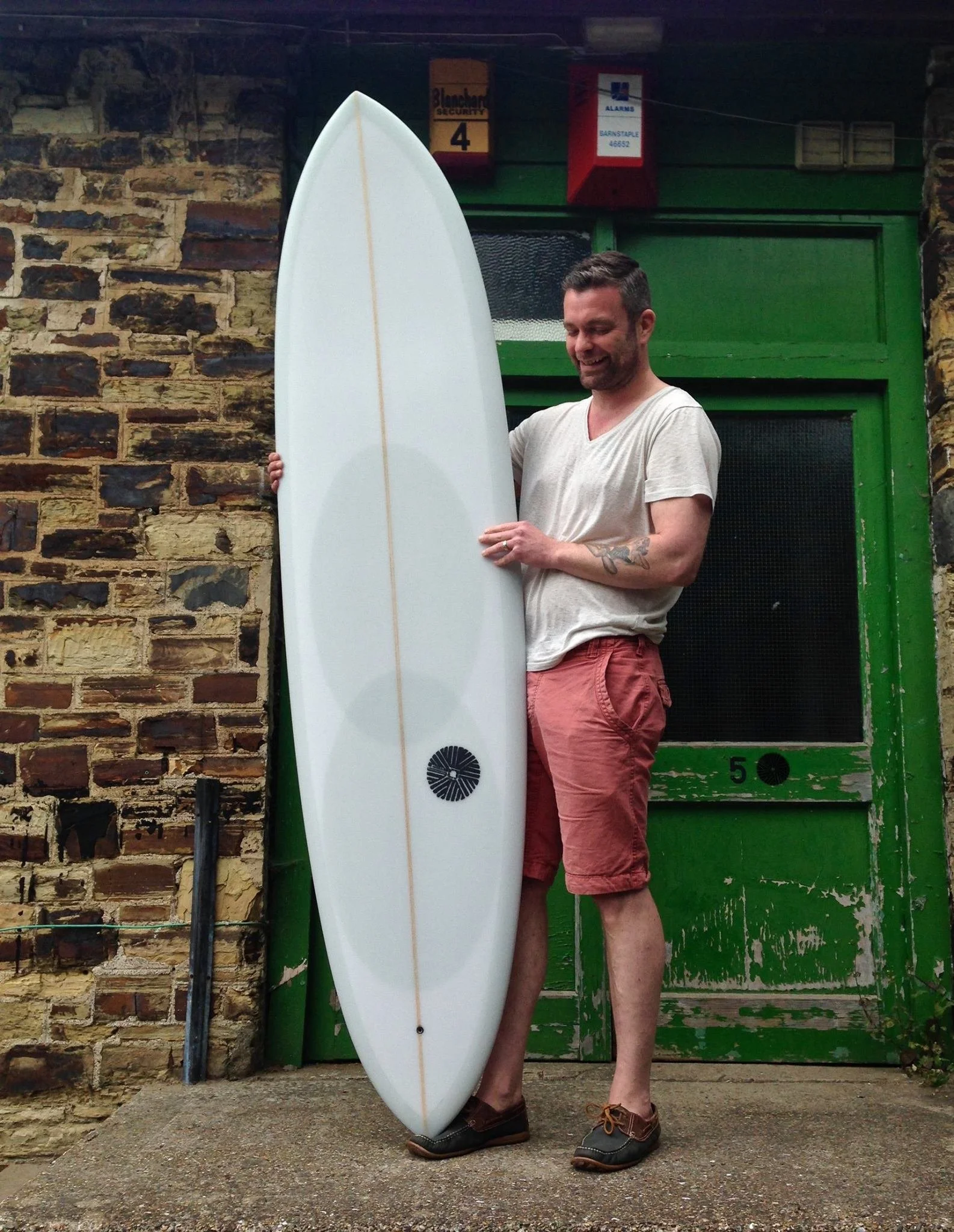 A man with a tattoo on his arm holding a white surfboard outdoors, standing against a brick wall and a green door, smiling and looking down.