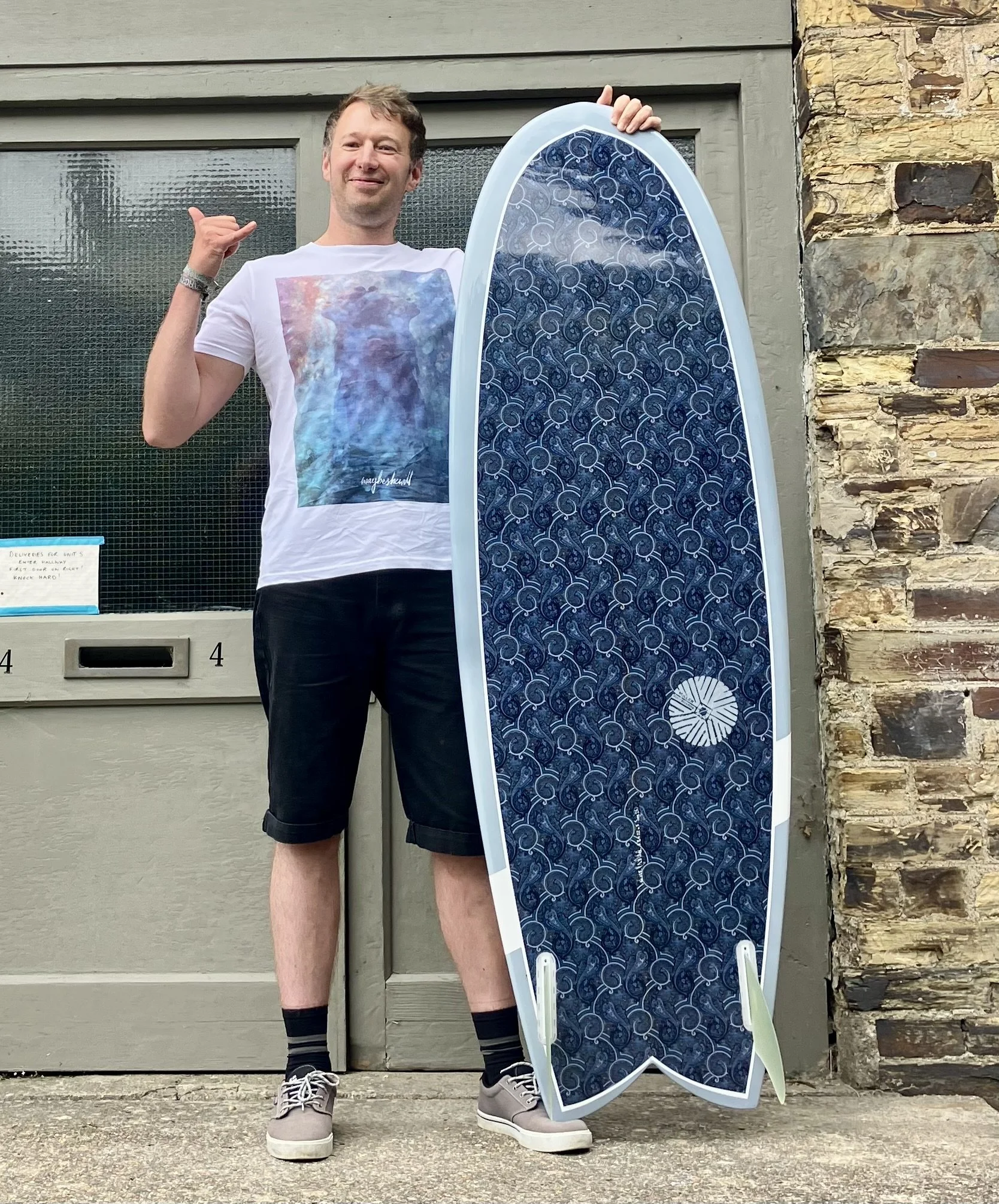 A man standing outside near a stone wall, holding a large surfboard with a blue patterned covering, and making a shaka sign with his right hand. He is wearing a white graphic t-shirt, black shorts, striped socks, and sneakers.