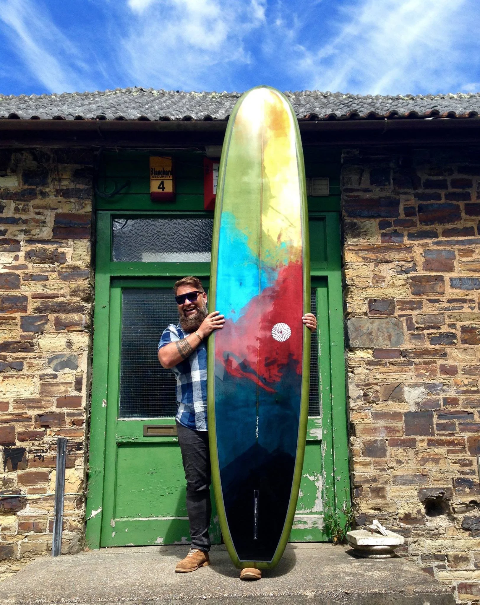 A man with a beard, sunglasses, and a plaid shirt stands outside a brick building with a green door, holding a colorful surfboard with a rainbow pattern.