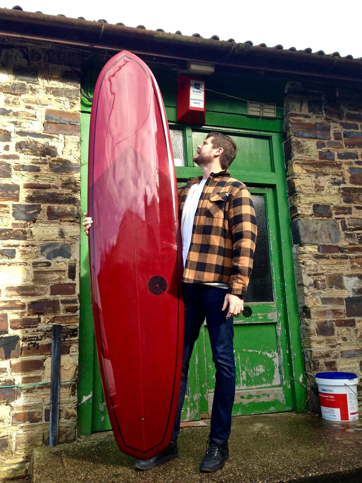 A man with a beard and short hair stands outside a green wooden door and brick wall, holding a red surfboard vertically. He is wearing a brown plaid jacket over a white shirt and dark jeans.