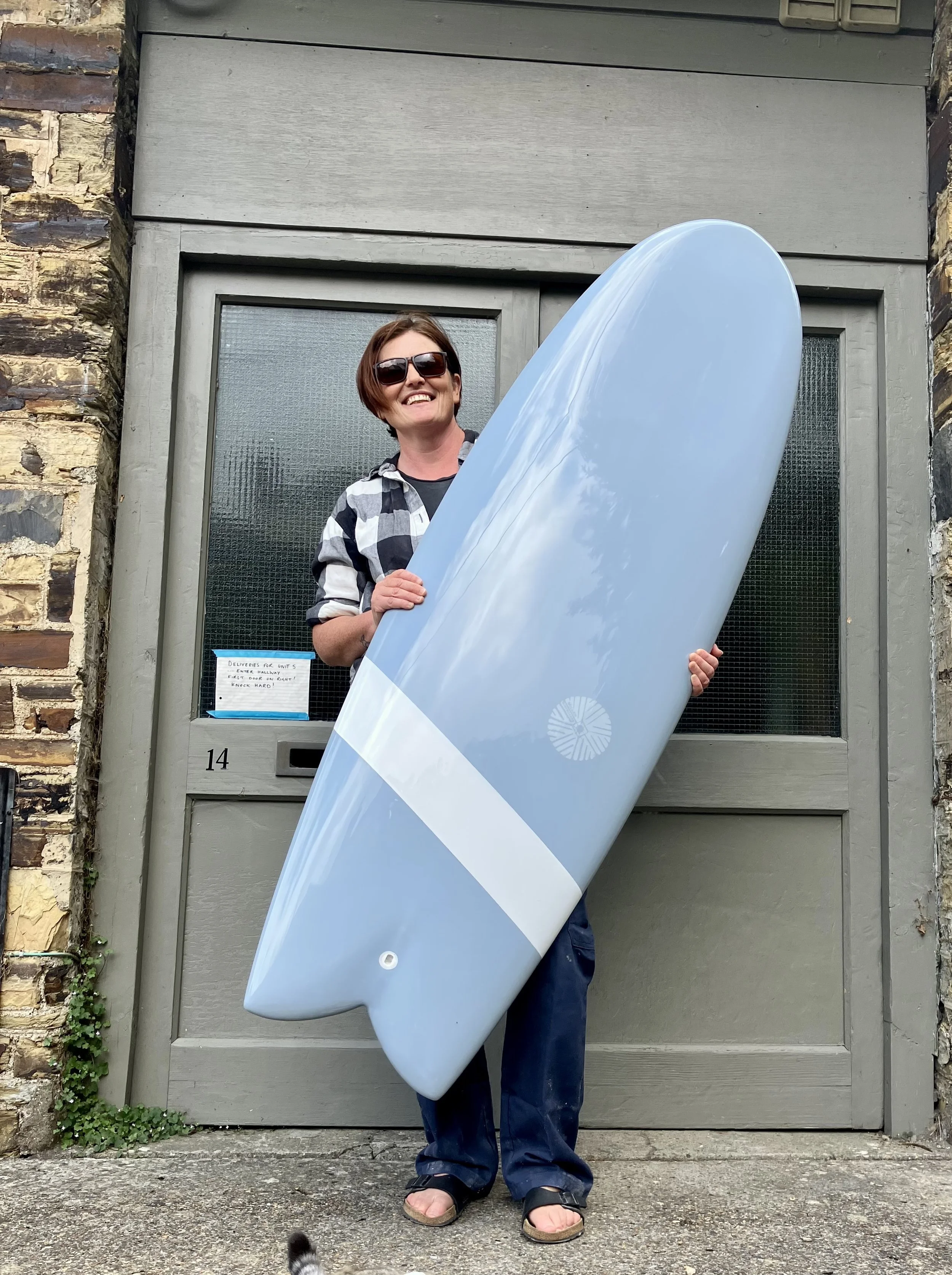 A smiling woman wearing sunglasses and a checkered shirt holds a large surfboard in front of a grey door and brick wall.