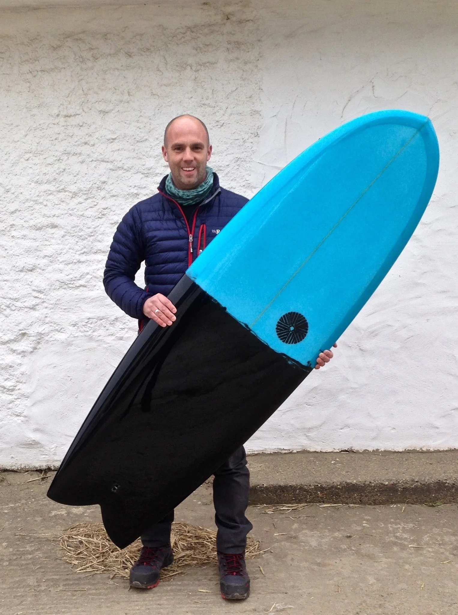 A man holding a large black and blue surfboard, standing on a dirt ground in front of a white wall.