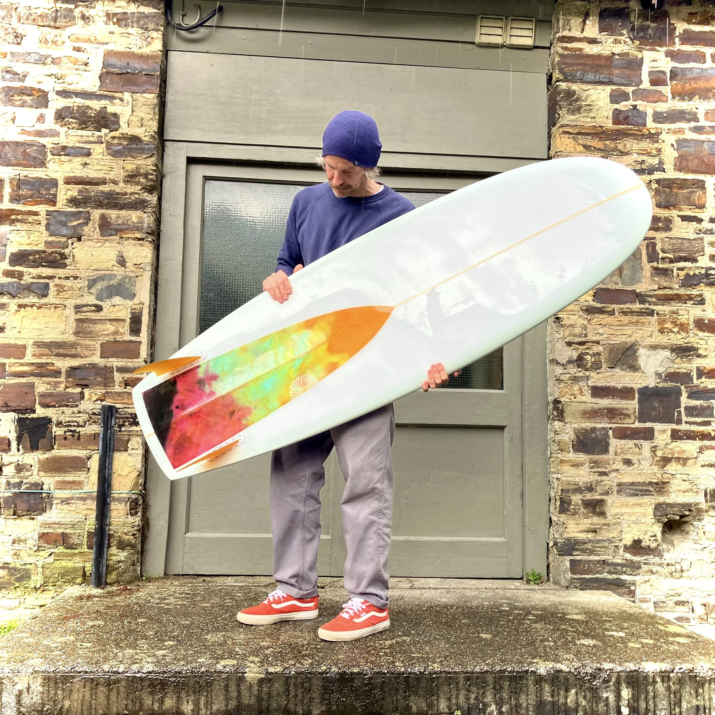 A man with a blue beanie, blue sweatshirt, gray pants, and red sneakers holding a surfboard with colorful designs against a brick wall background.
