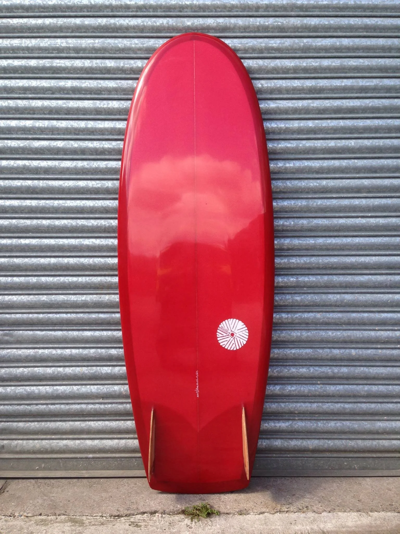 Red surfboard with a circular white logo, leaning against a gray corrugated metal wall.