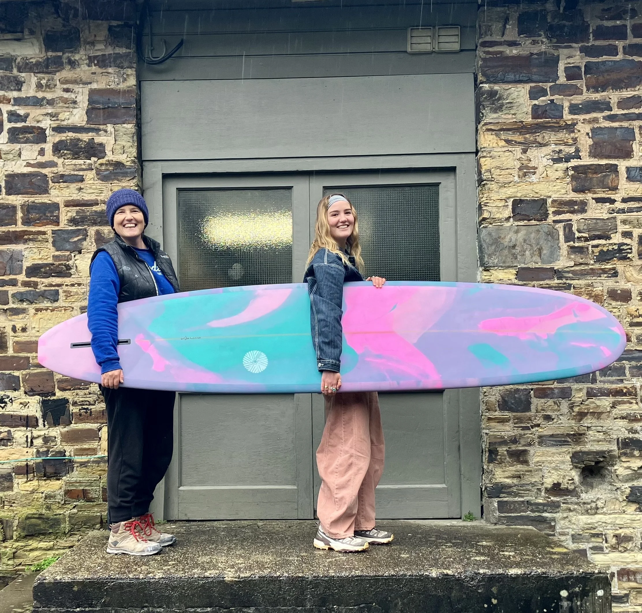 Two women standing outside in front of a stone wall and a door, holding a colorful surfboard. One woman is wearing a blue beanie, black vest, and athletic shoes, and the other is wearing a denim jacket, pink pants, and sneakers. Both are smiling and 