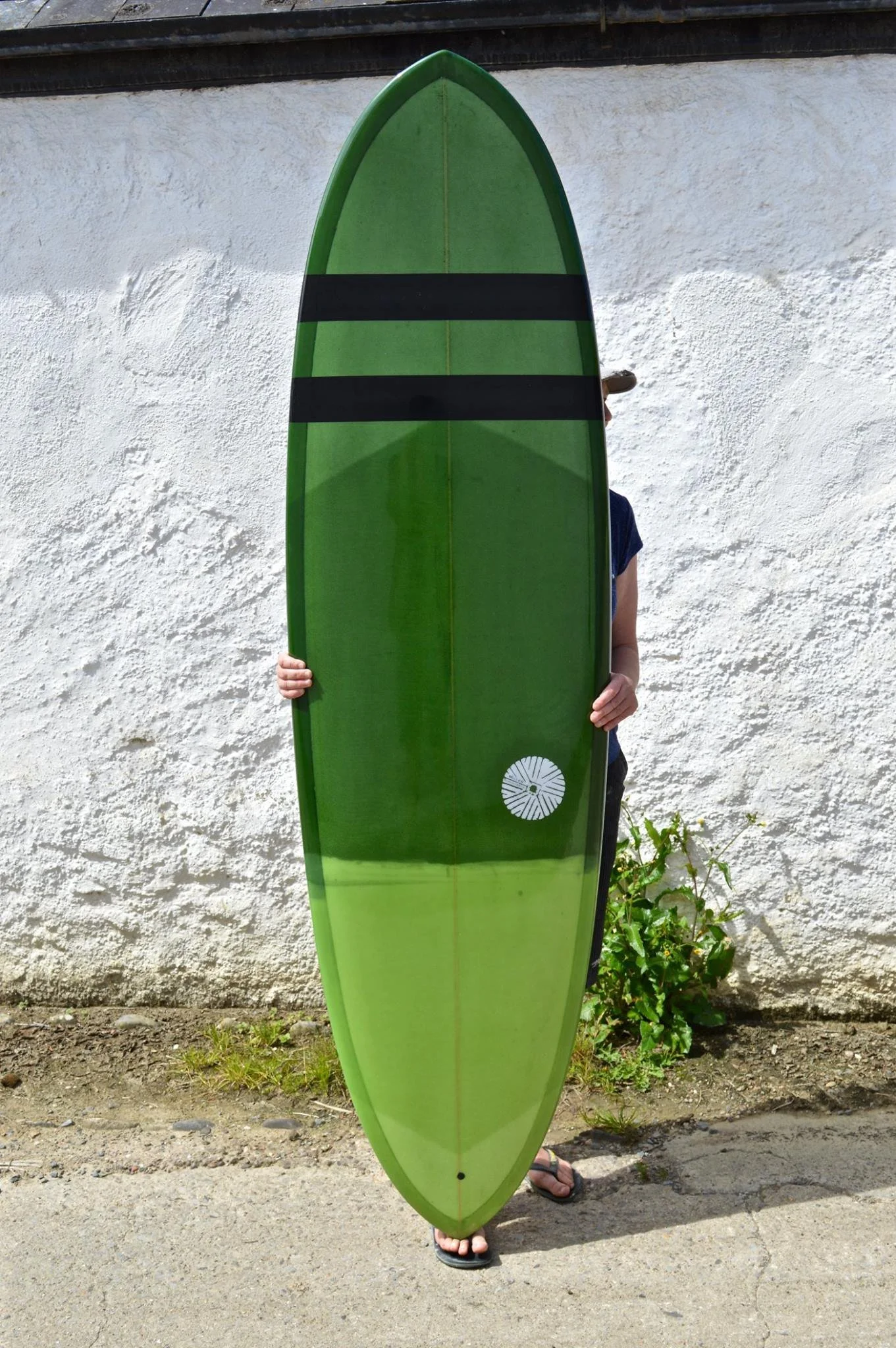 Person holding a large green surfboard outdoors, standing on pavement in front of a white textured wall, wearing flip-flops.