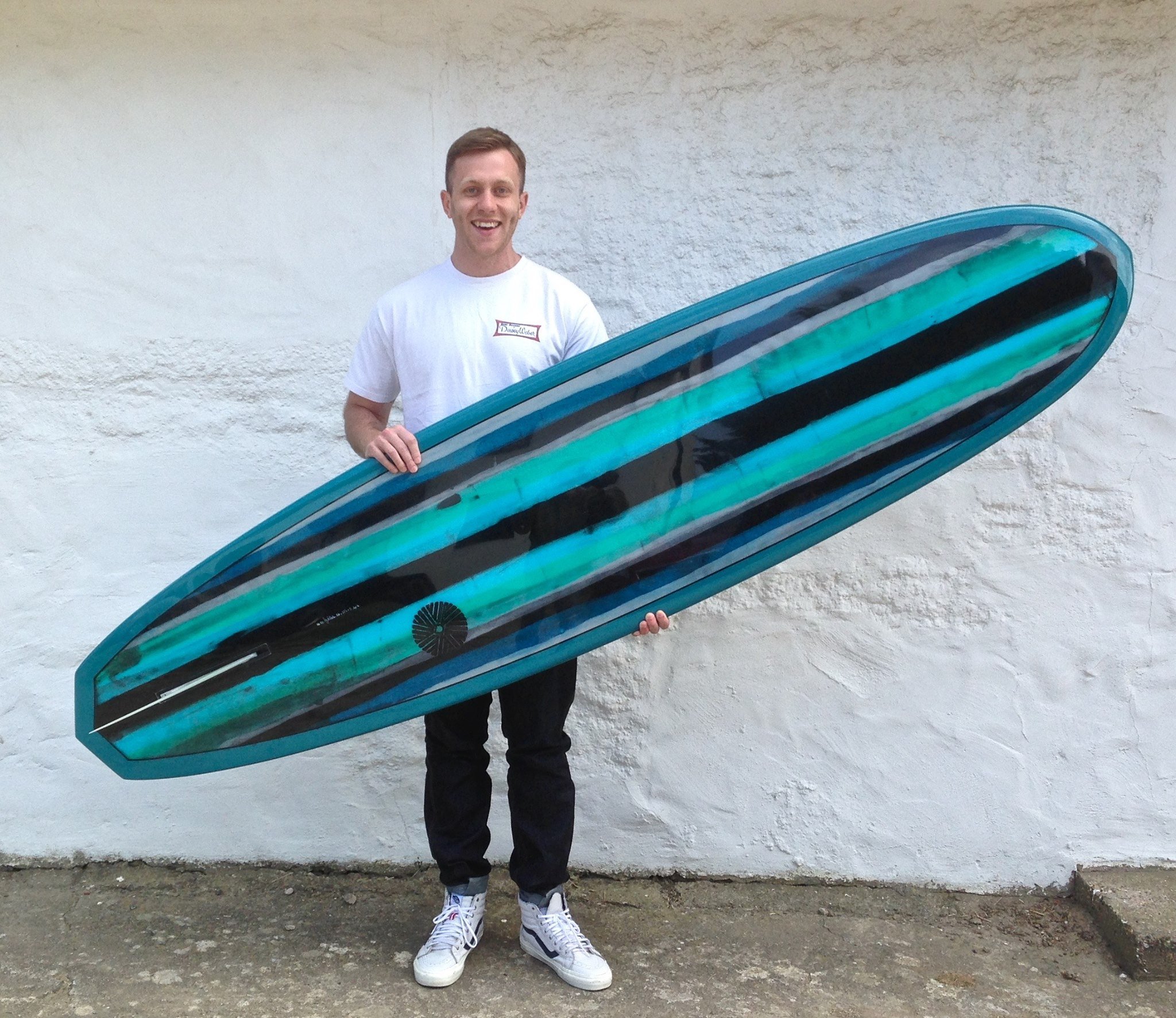 A man standing outdoors holding a large, colorful surfboard in front of a white textured wall.