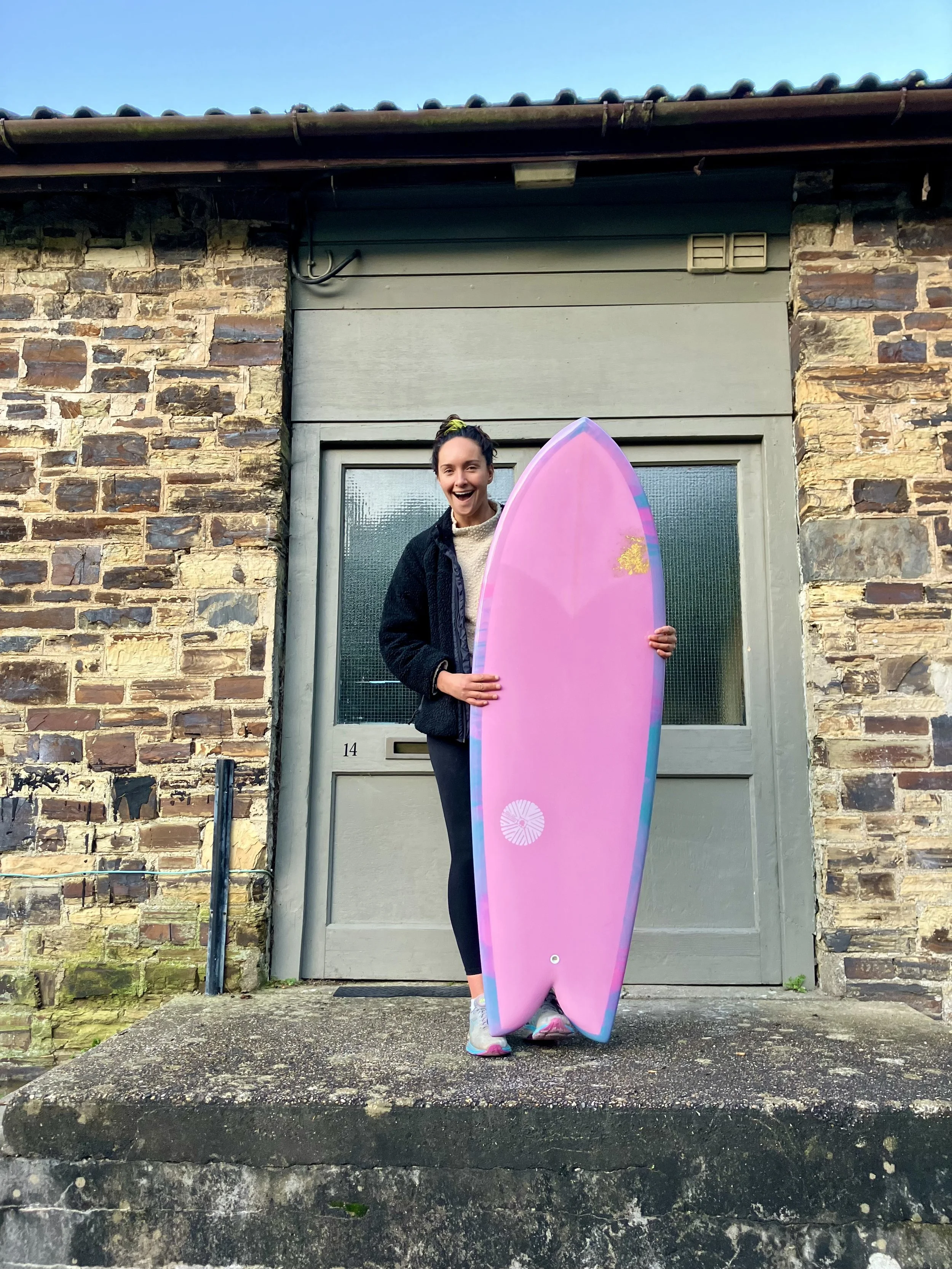 A woman standing on a concrete step outside a brick house, holding a pink surfboard with a smile on her face.