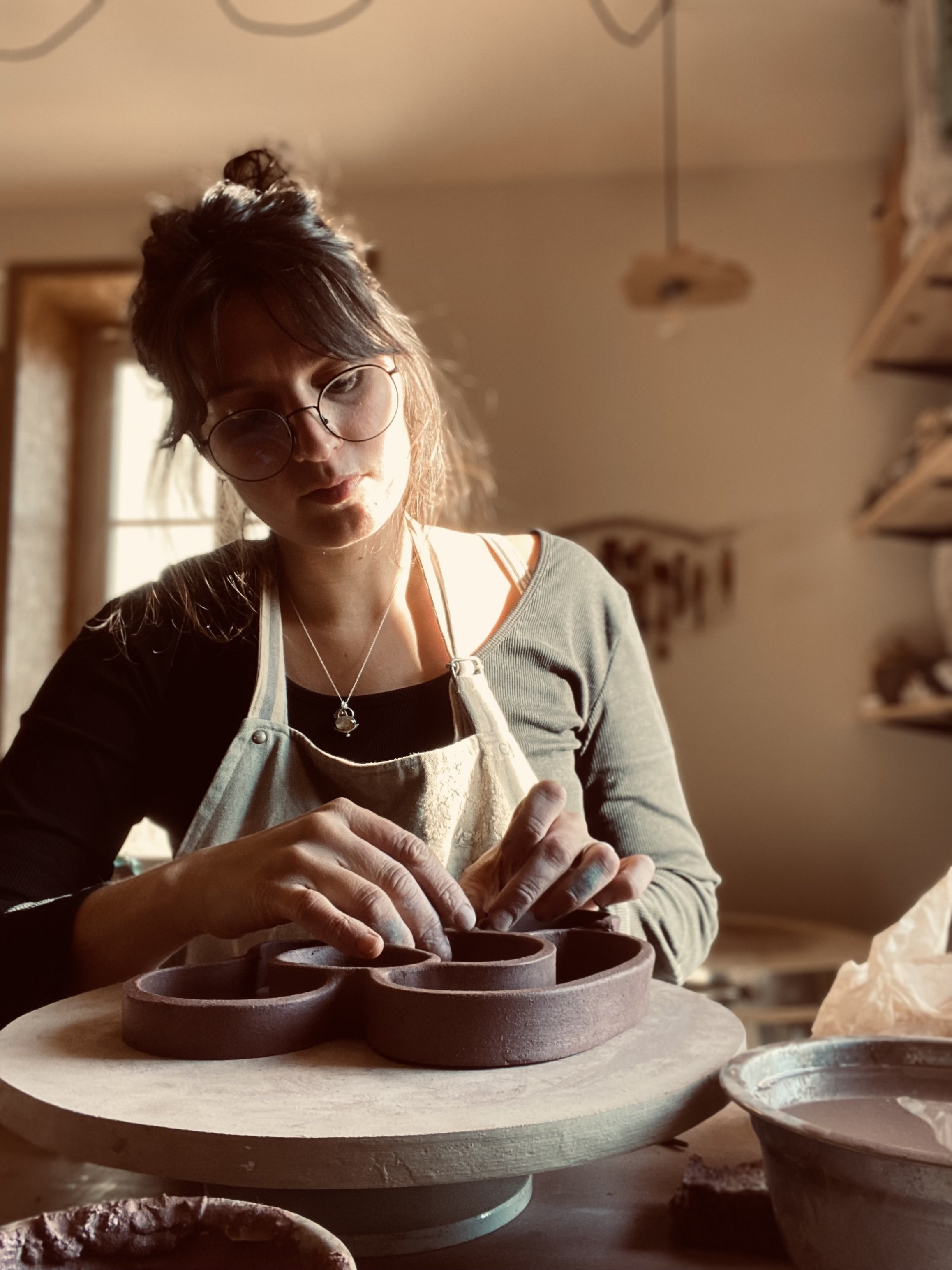 Jeune femme portant des lunettes et un tablier en train de sculpter de la céramique dans un atelier lumineux.