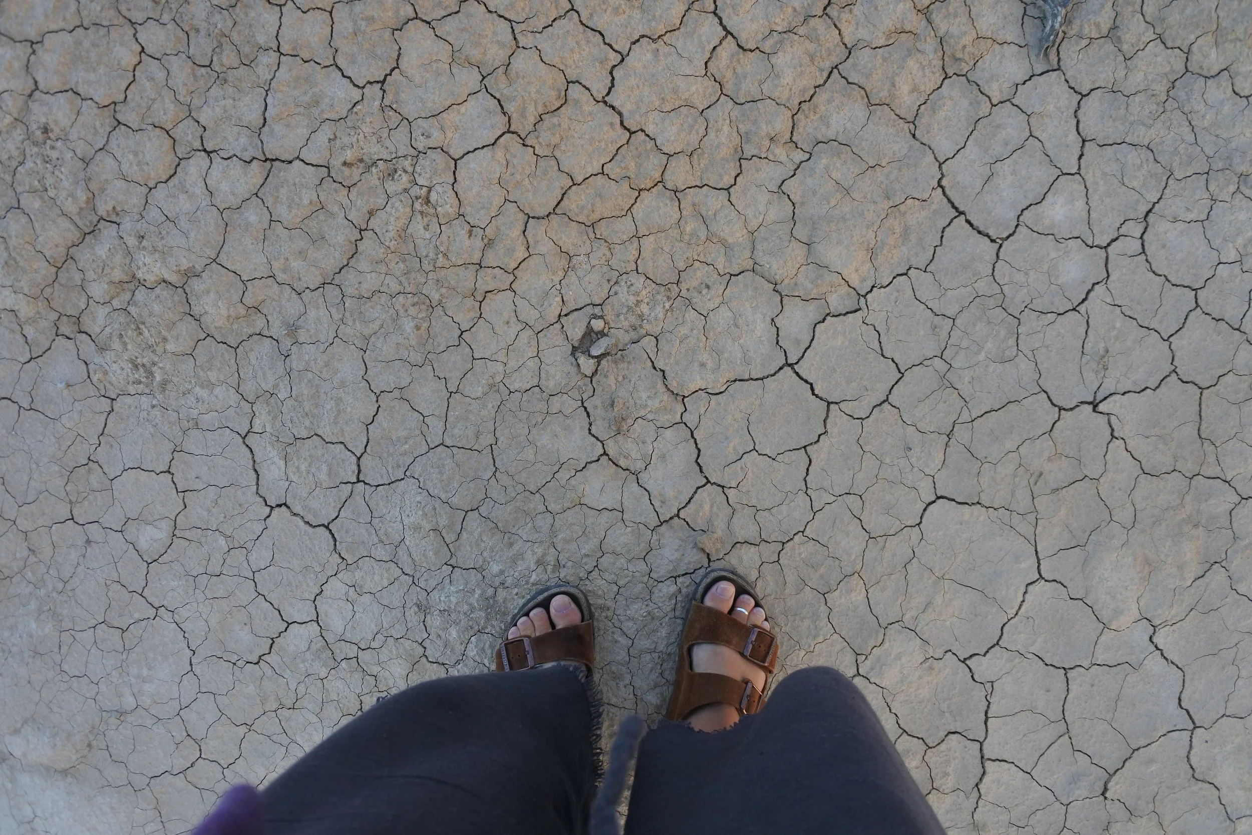 Photo prise depuis le point de vue d'une personne portant des sandales en cuir marron, sur un sol craquelé sec et aride.