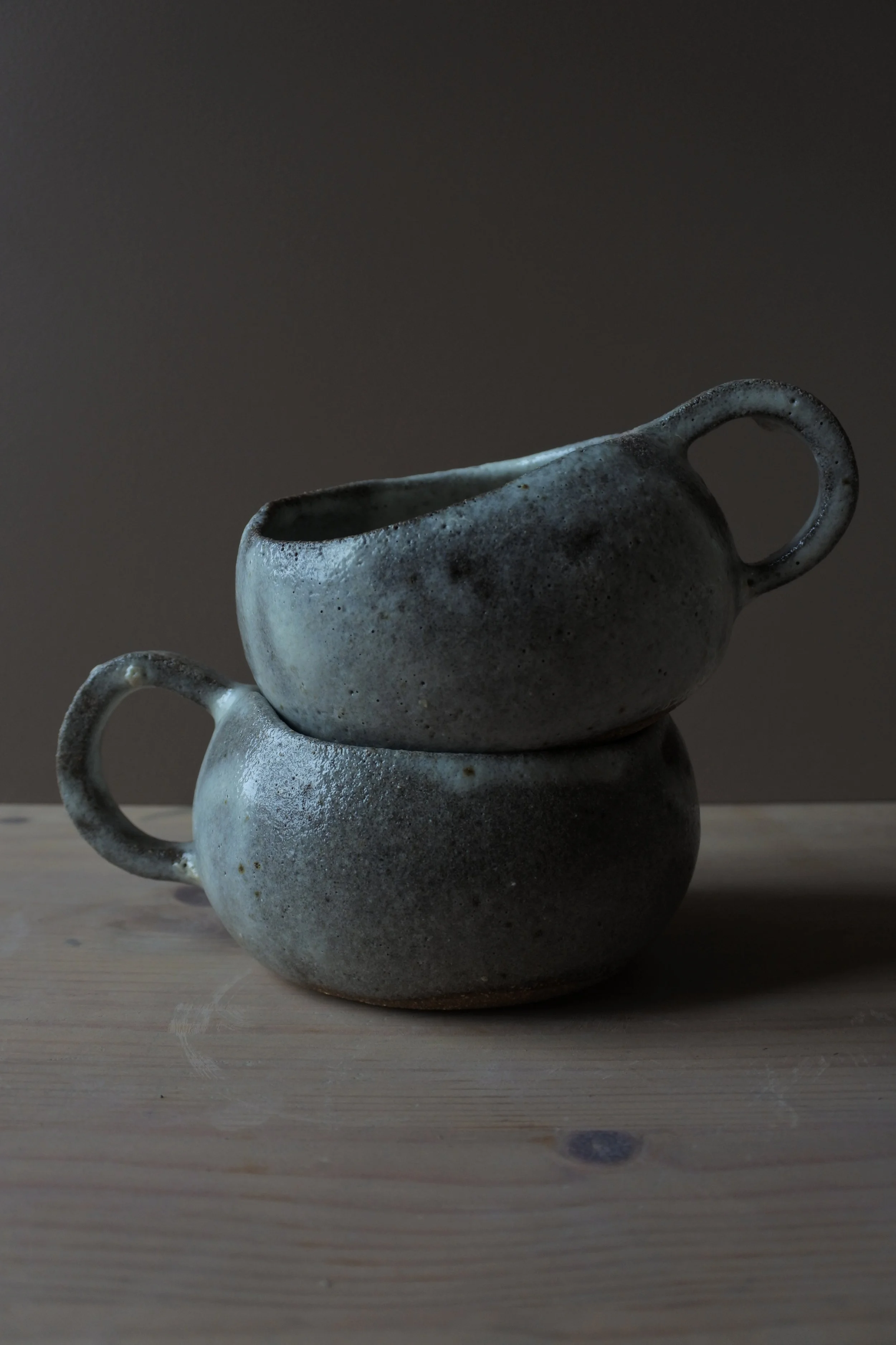 Deux mug en céramique gris disposés l'un sur l'autre sur une surface en bois, avec un fond sombre.
