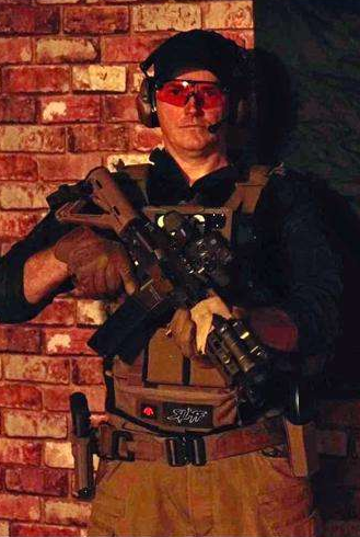 A man dressed in tactical gear, wearing headphones, red safety glasses, and camouflage gloves, holding a firearm and standing against a brick wall.