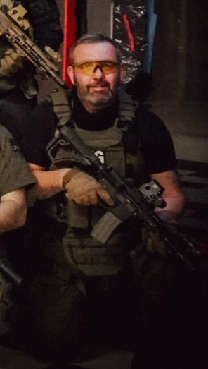 A man with a beard wearing tactical gear, including yellow eye protection, holding a rifle indoors.
