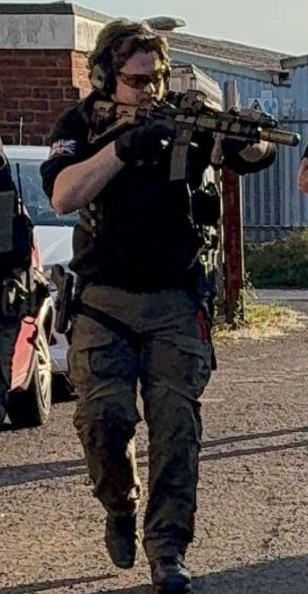Person wearing black tactical gear, sunglasses, and headphones, aiming a rifle outdoors.