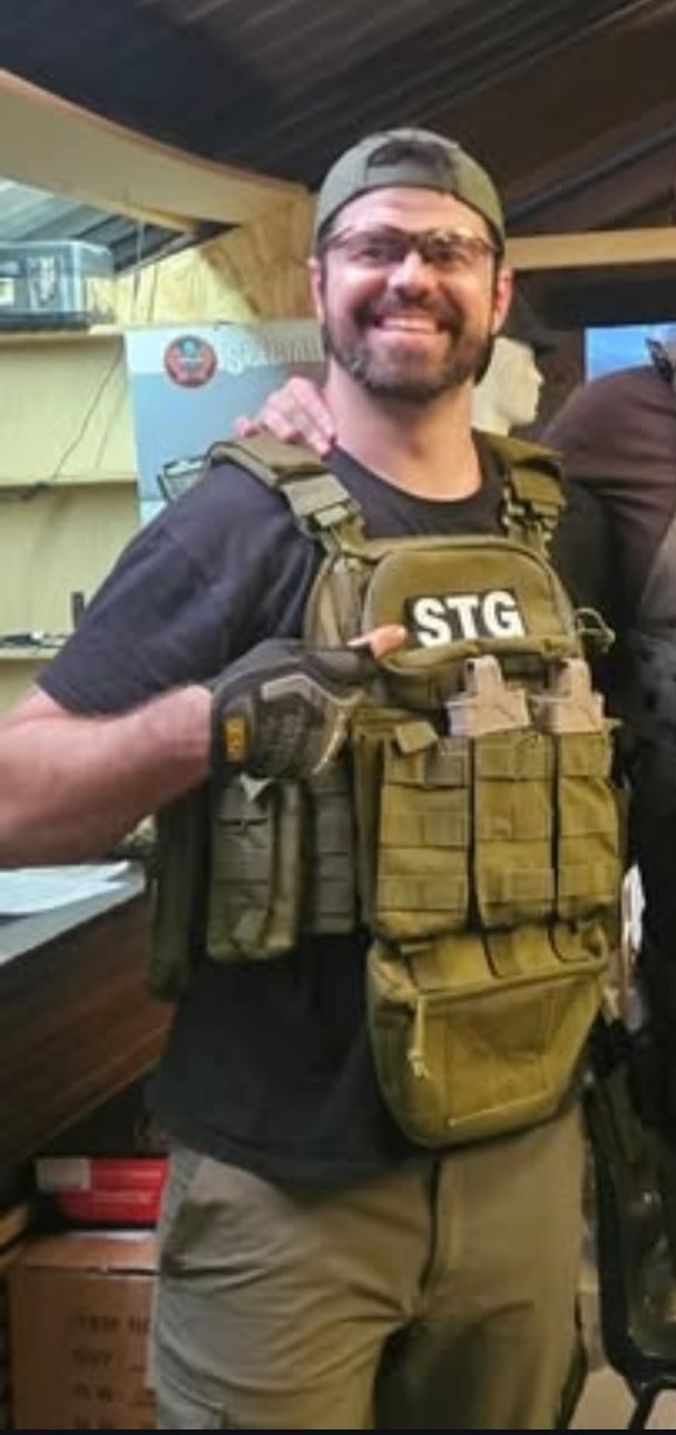 A man wearing glasses, a backwards baseball cap, a black T-shirt, and tan tactical gear with an 'STG' patch, smiling in an indoor setting with wooden walls and shelves.