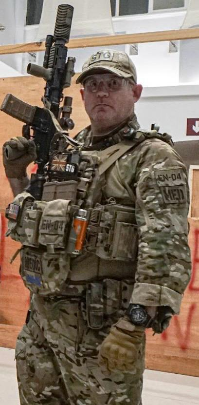 A soldier in camouflage uniform with tactical gear holding a rifle, standing indoors.