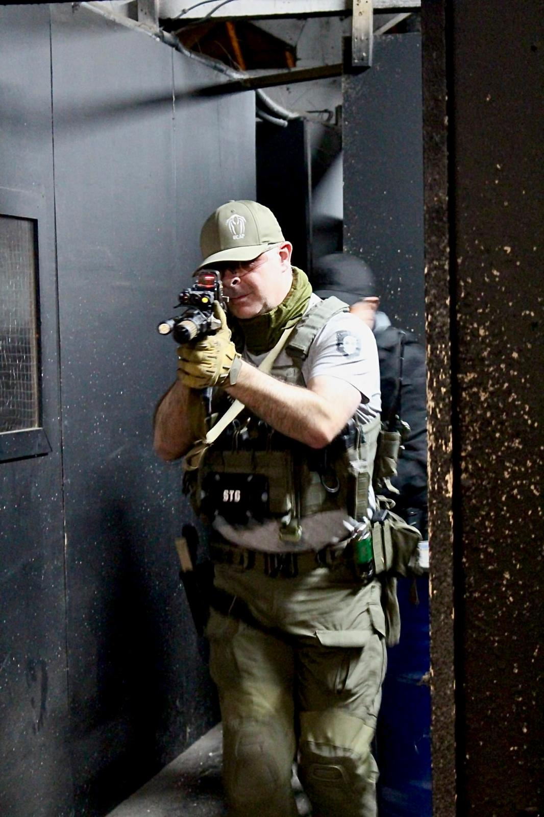 A man in tactical gear aims a rifle around a corner in a dimly lit indoor setting.