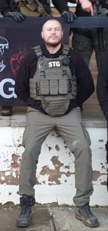 A man wearing tactical gear and a black hoodie, sitting on a ledge outdoors, with a group of people in the background.
