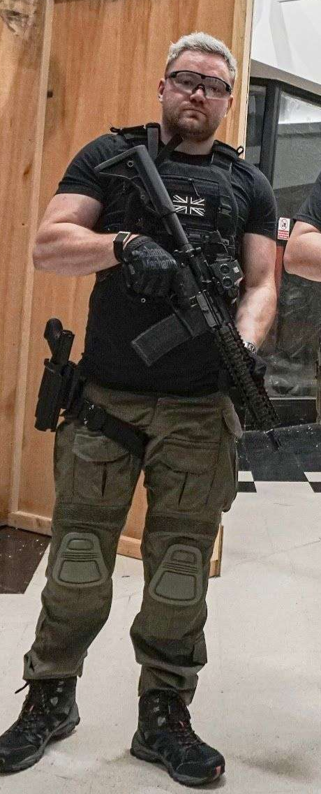 Man dressed in tactical gear holding a rifle, wearing glasses, black t-shirt with a Union Jack patch, camouflage pants, gloves, and boots, standing indoors against a wooden wall.