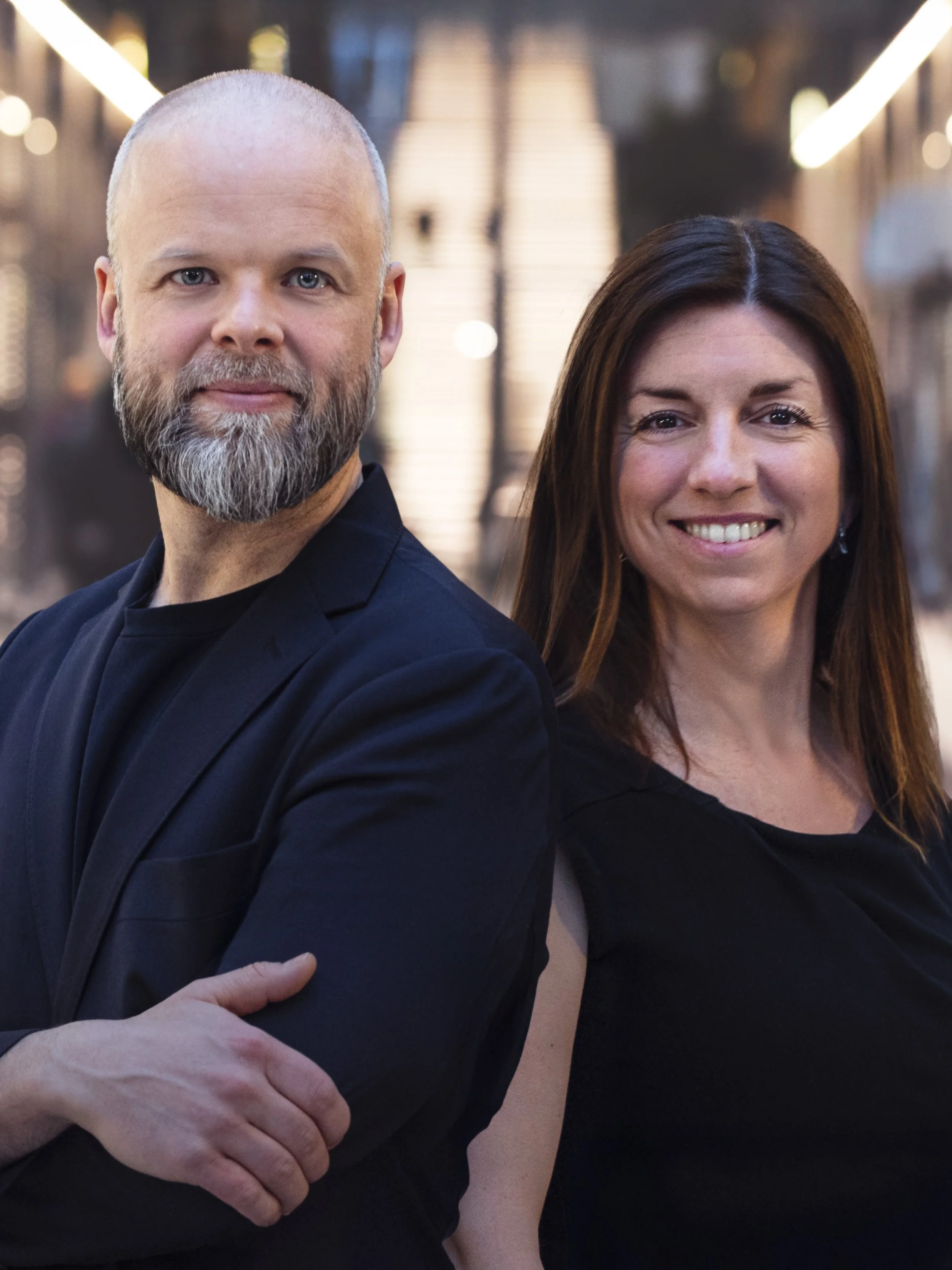 Two people standing close together outdoors at night, smiling and looking at the camera. The man has a bald head and a beard, wearing a dark suit. The woman has long brown hair and is wearing a black top.