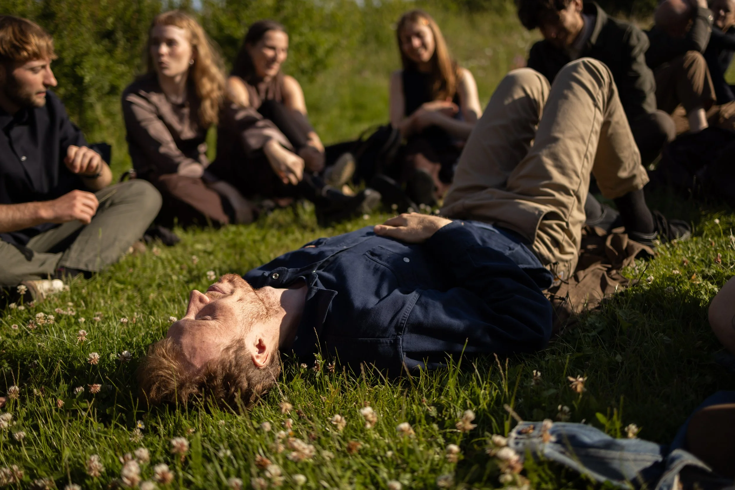 A man with red hair lies on the grass with his eyes closed, while a group of young adults sit nearby on the grass, engaging in conversation in a park during daytime.