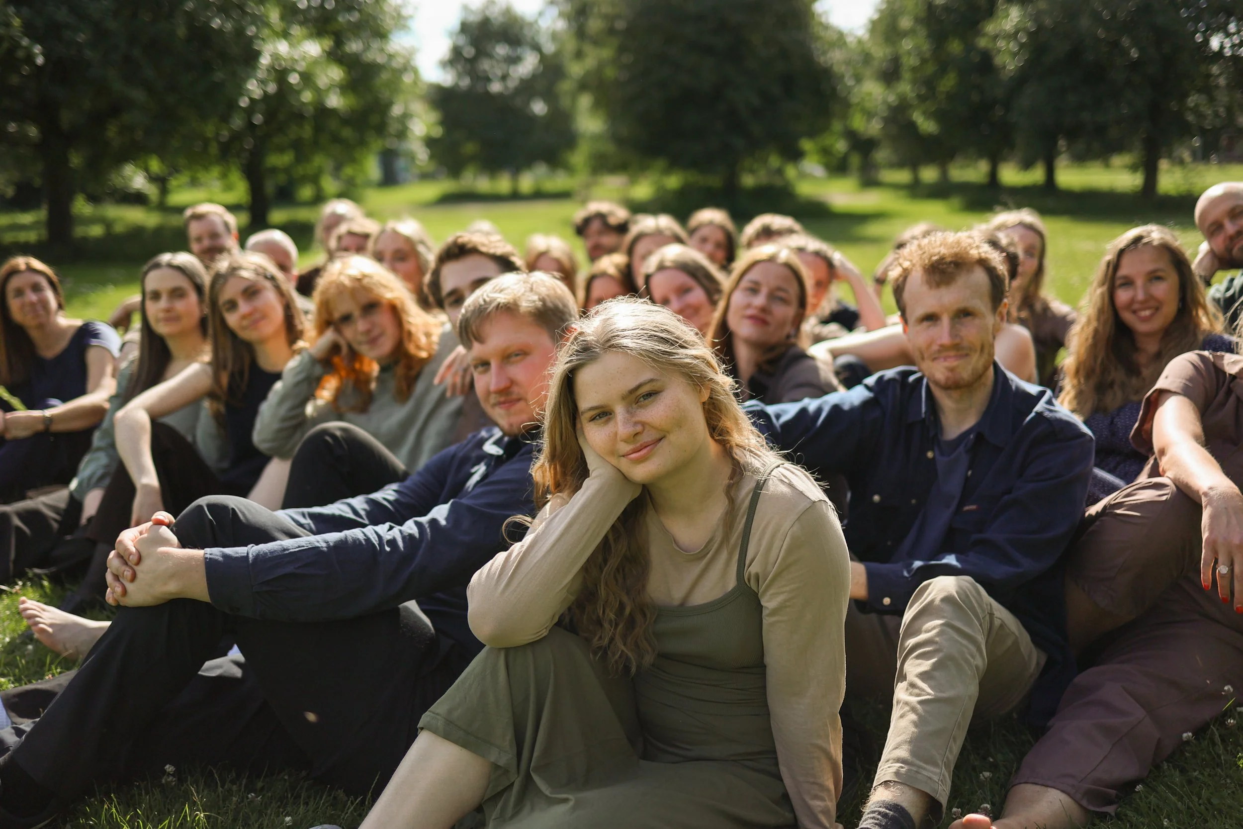 A group of people sitting on grass outdoors in a park, smiling and relaxing on a sunny day.