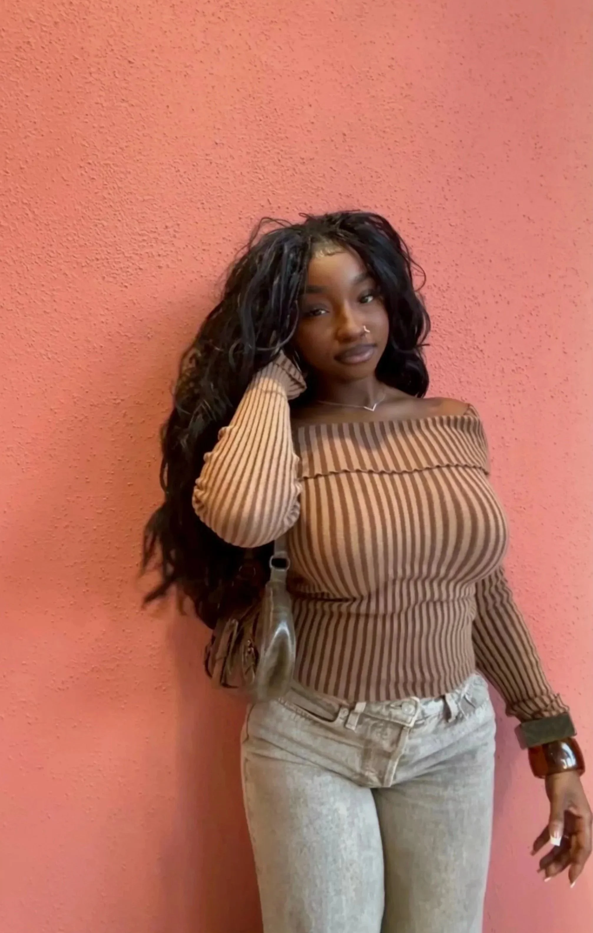 A young woman with long curly black hair standing against a pink wall, wearing a brown and beige striped off-shoulder top and light-colored jeans.