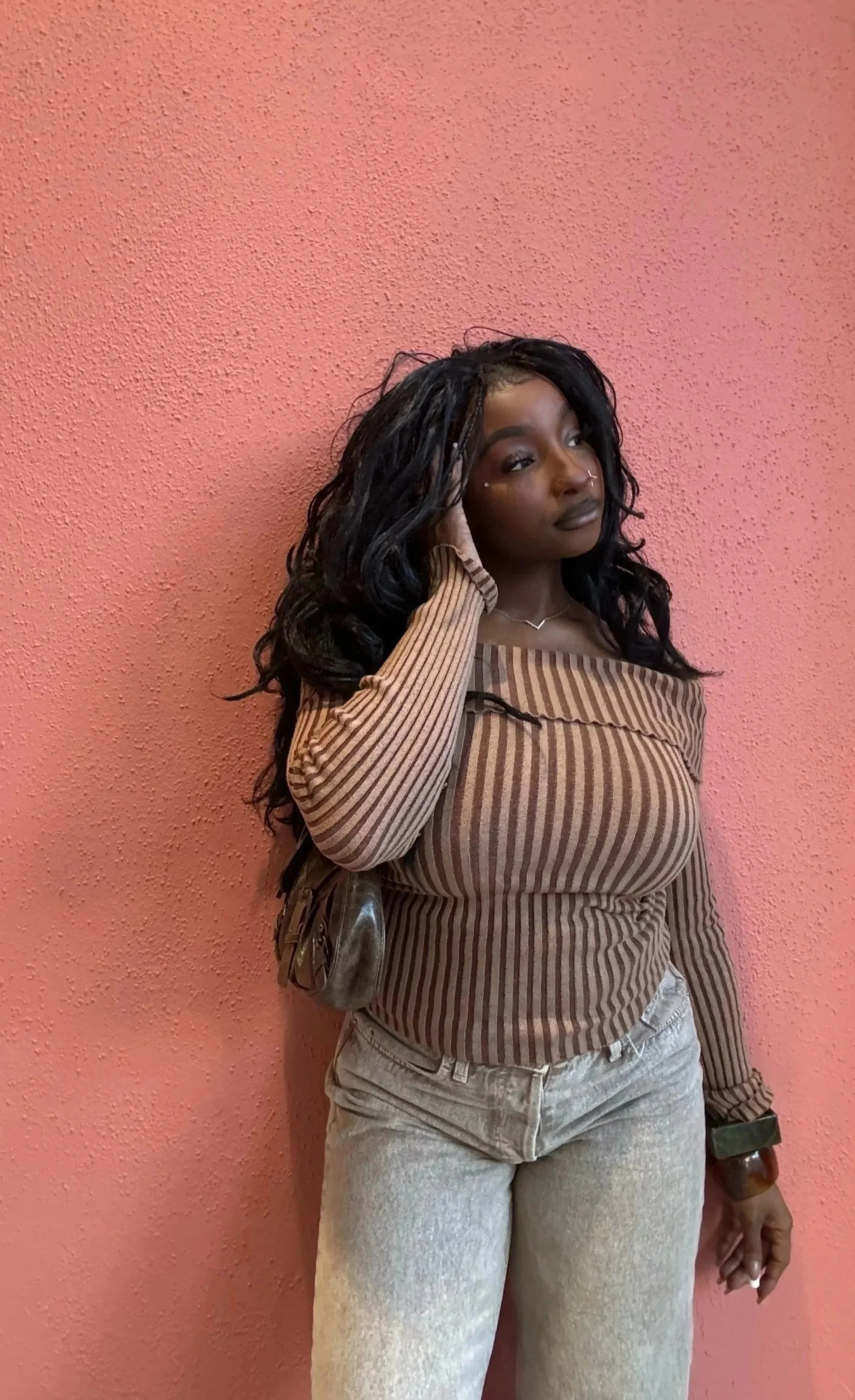 A young woman with long dark curly hair stands against a pink textured wall, wearing a brown and tan striped off-the-shoulder top and light-colored jeans, touching her hair with one hand and holding an object in her other hand.