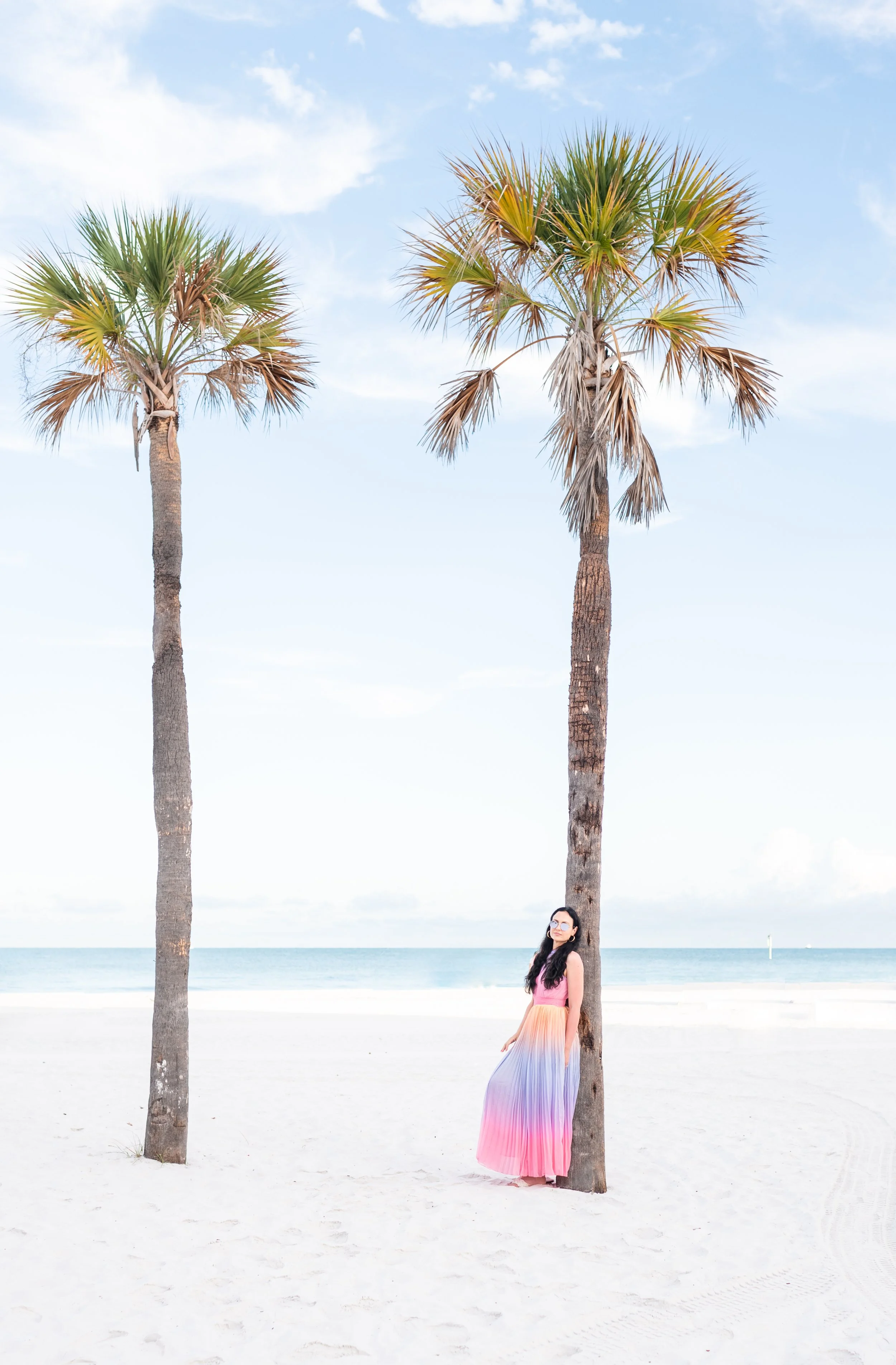 A woman in a multicolored dress stands next to a palm tree on a sandy beach with the ocean in the background and a blue sky with some clouds.