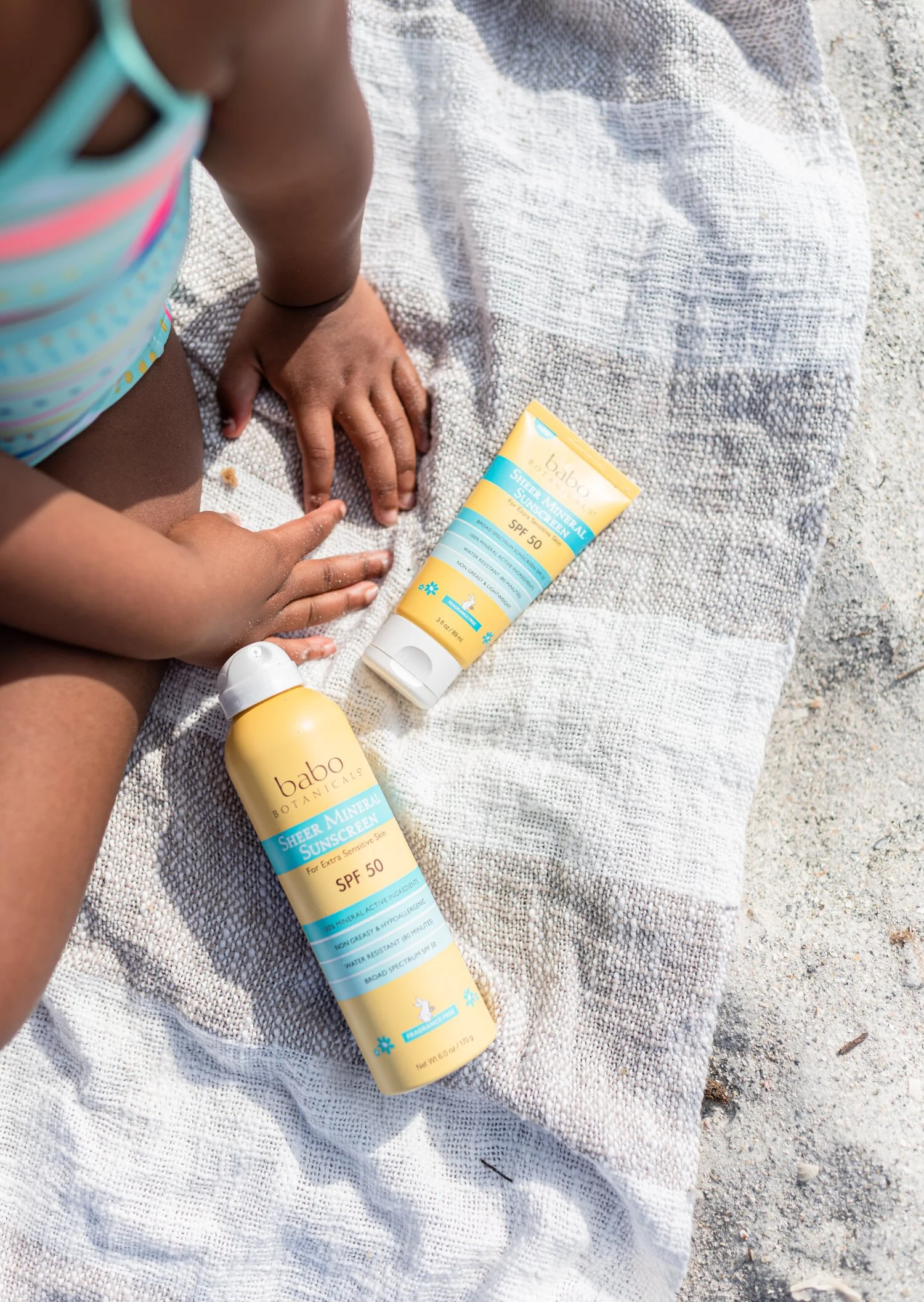 A child's hand and knee on a towel at the beach next to two bottles of Babo Botanicals SPF 50 sunscreen, one spray and one lotion.