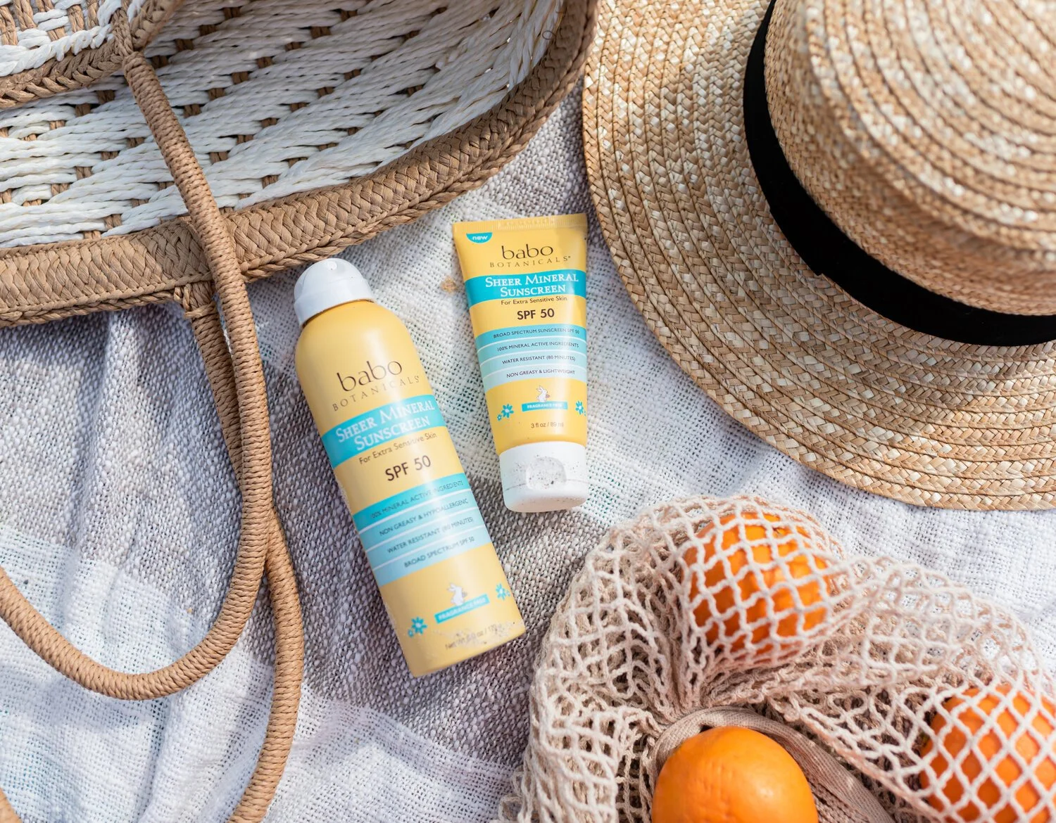 Two bottles of Babo Botanicals Sheer Mineral Sunscreen SPF 50, a straw hat with black band, a woven basket, a mesh bag with oranges, and woven bags on a textured cloth surface.