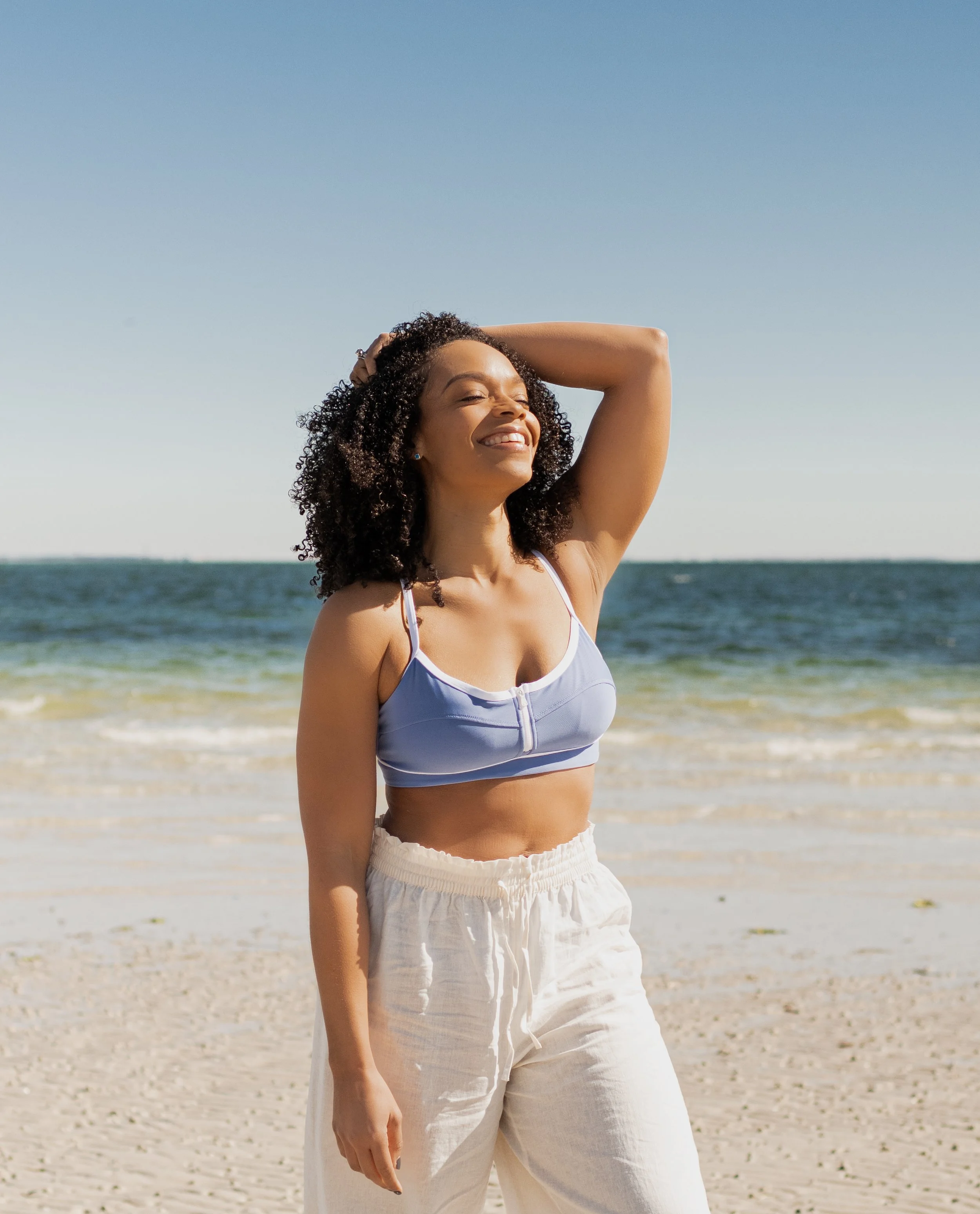 A woman standing on a sandy beach with her eyes closed, smiling, wearing a blue sports bra and white pants, with the ocean and clear sky in the background.