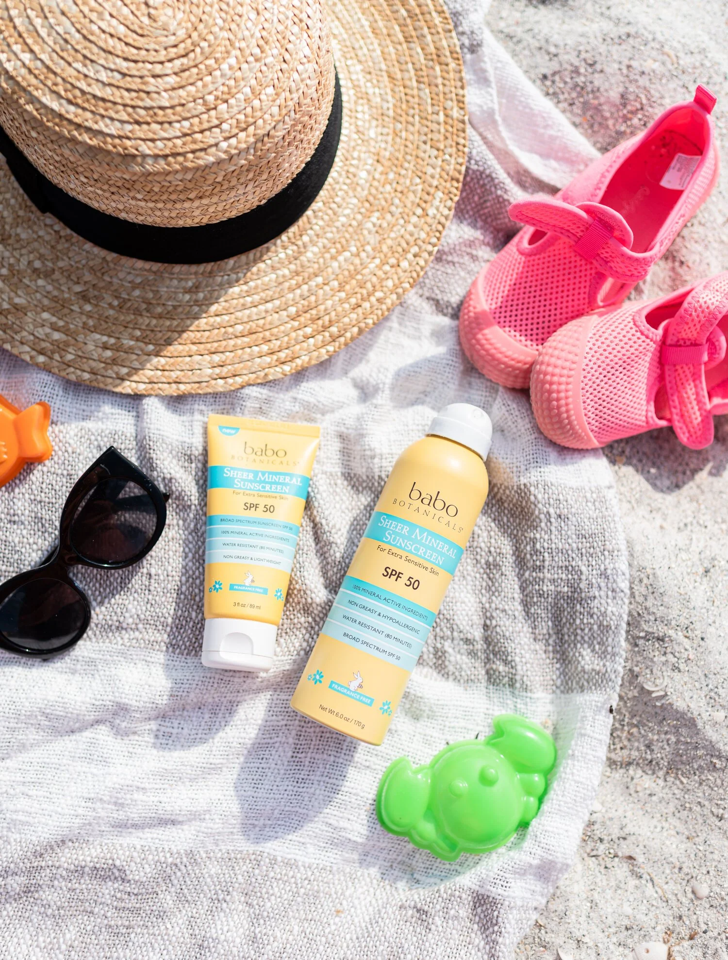 Beach scene with a straw hat, pink water shoes, sunglasses, sunscreen lotion, a green toy, and a cloth on sand.