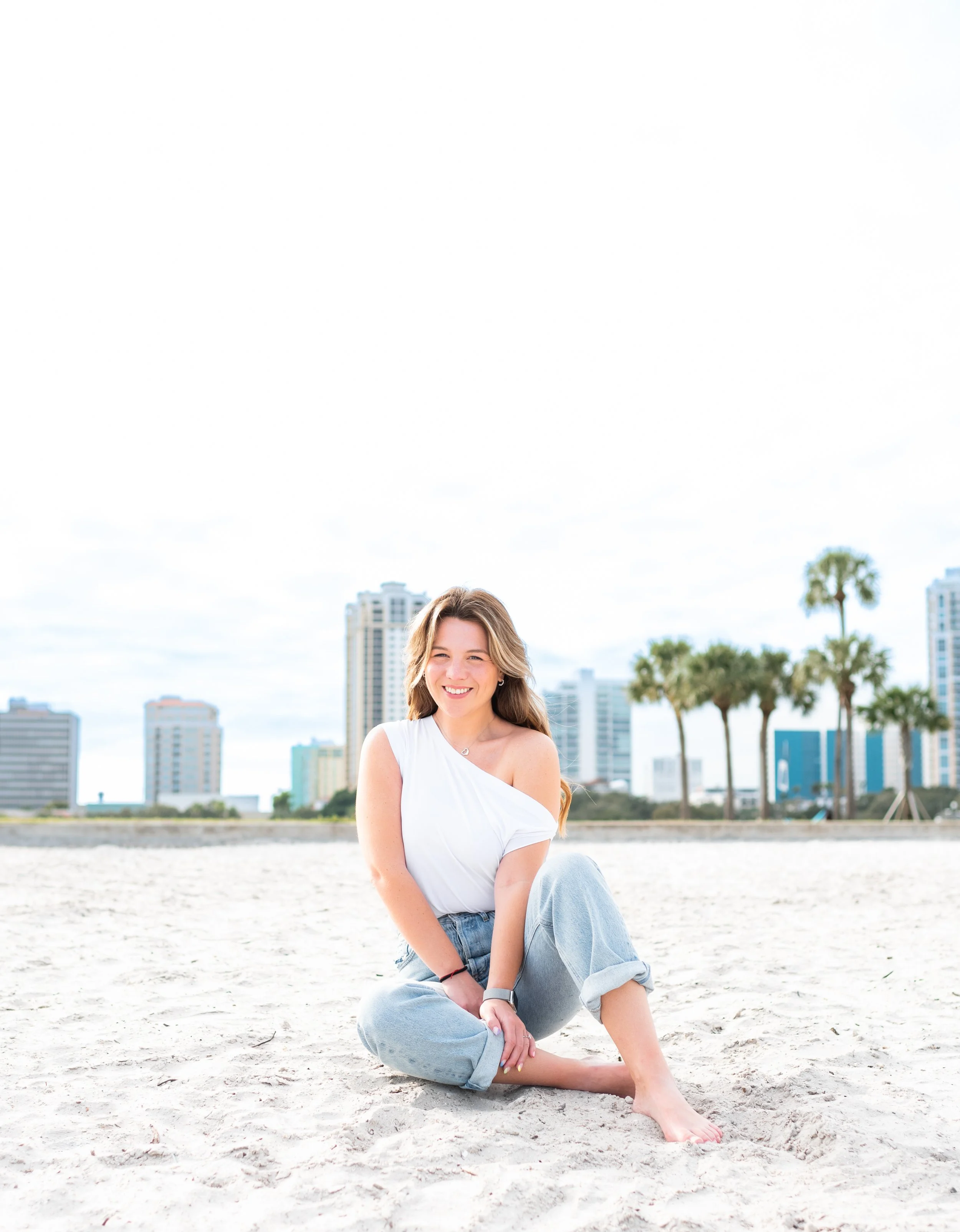 Young woman sitting on sandy beach with city skyline and palm trees in the background, smiling.