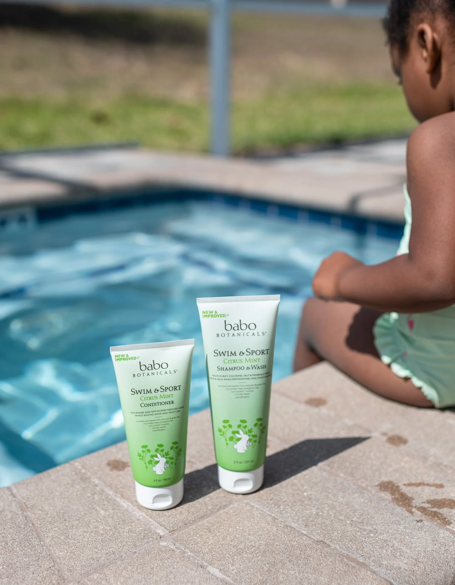 Two tubes of Babo Botanicals Swim & Sport Citrus Mint shampoo and conditioner placed on a poolside surface, with a young girl sitting by the pool in the background.