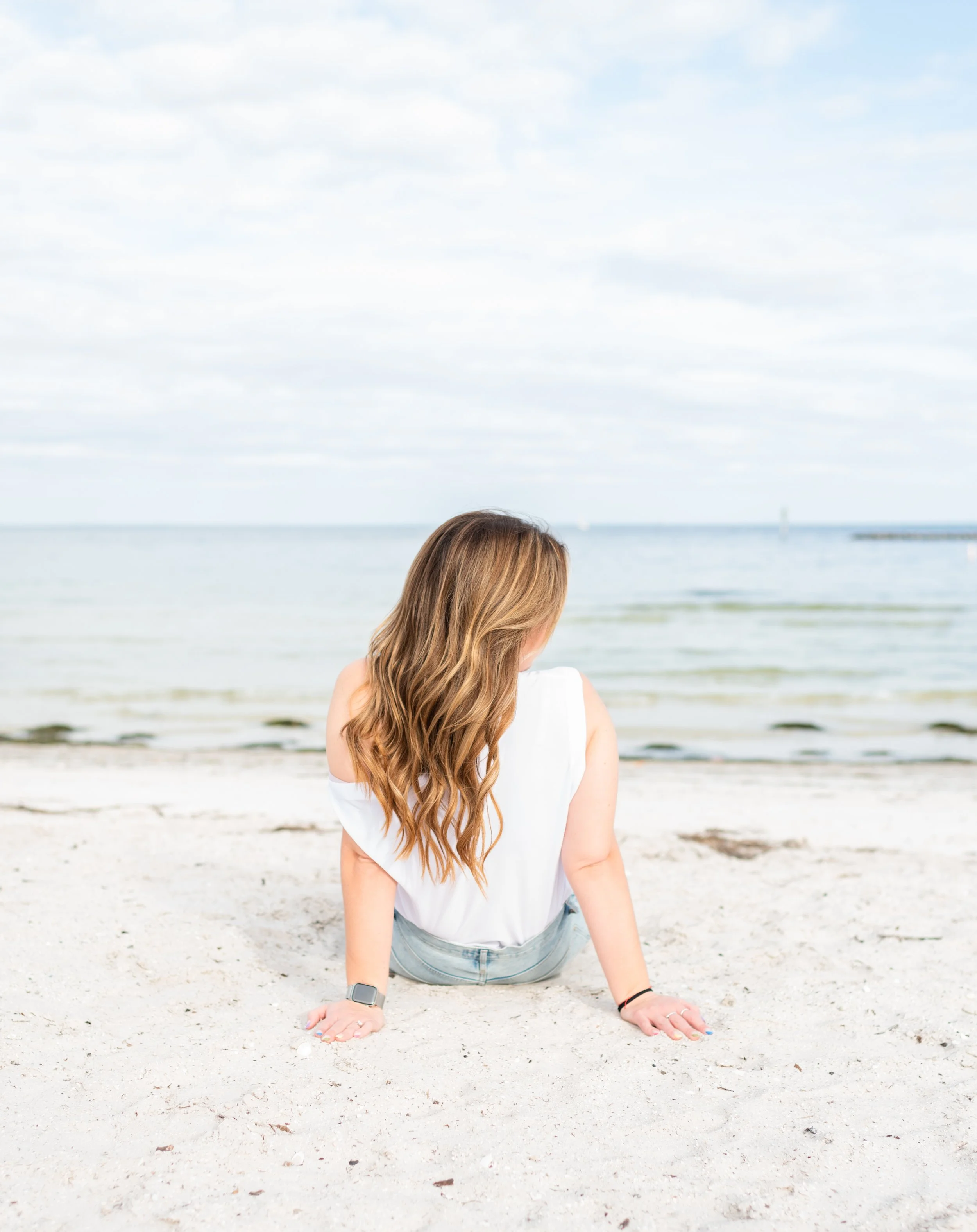 A woman with long wavy hair sitting on a sandy beach facing the calm ocean, wearing a white sleeveless top and denim shorts.