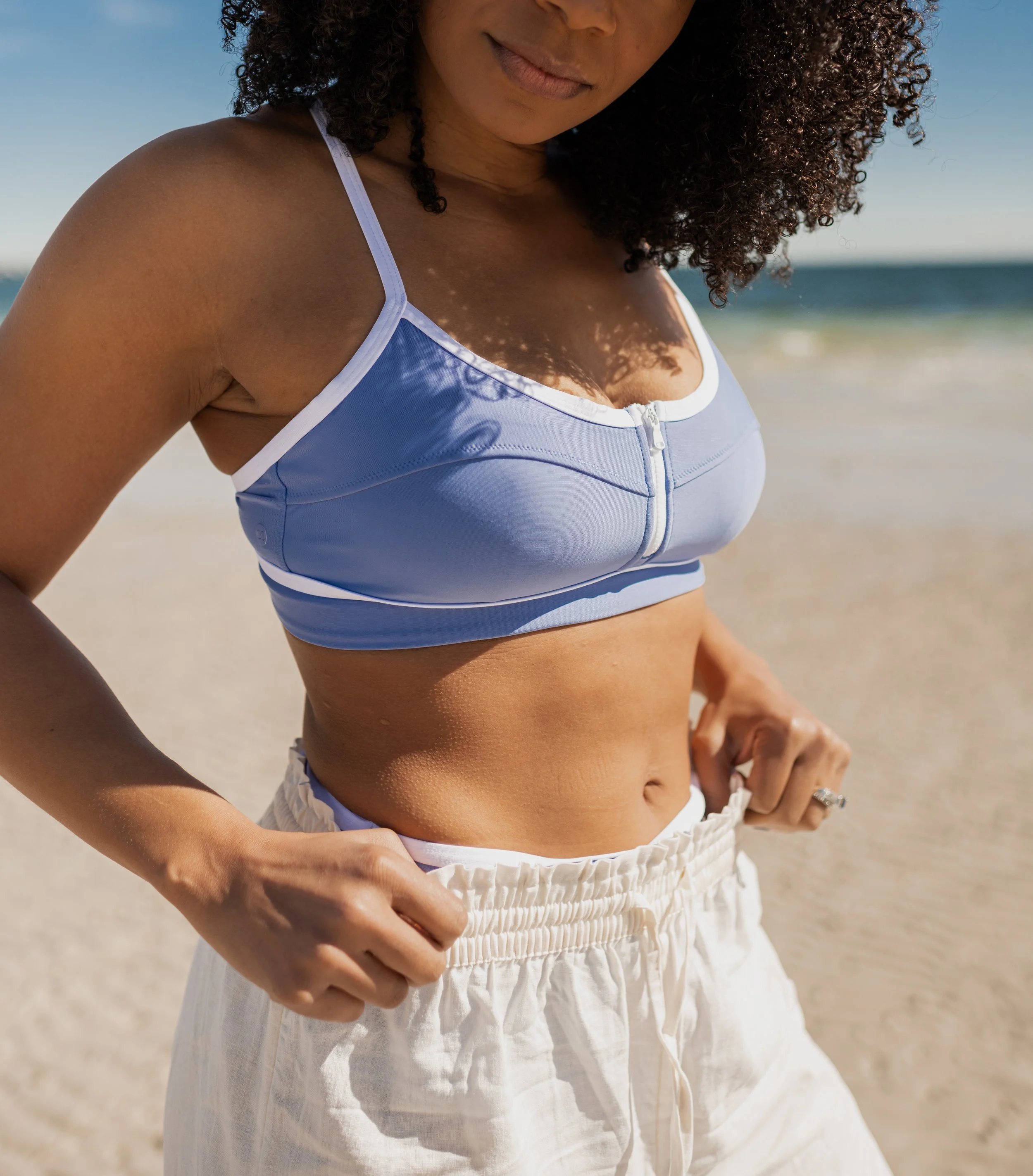 A woman on a beach adjusting her white shorts, wearing a blue zip-up sports bra, with the ocean and sky in the background.