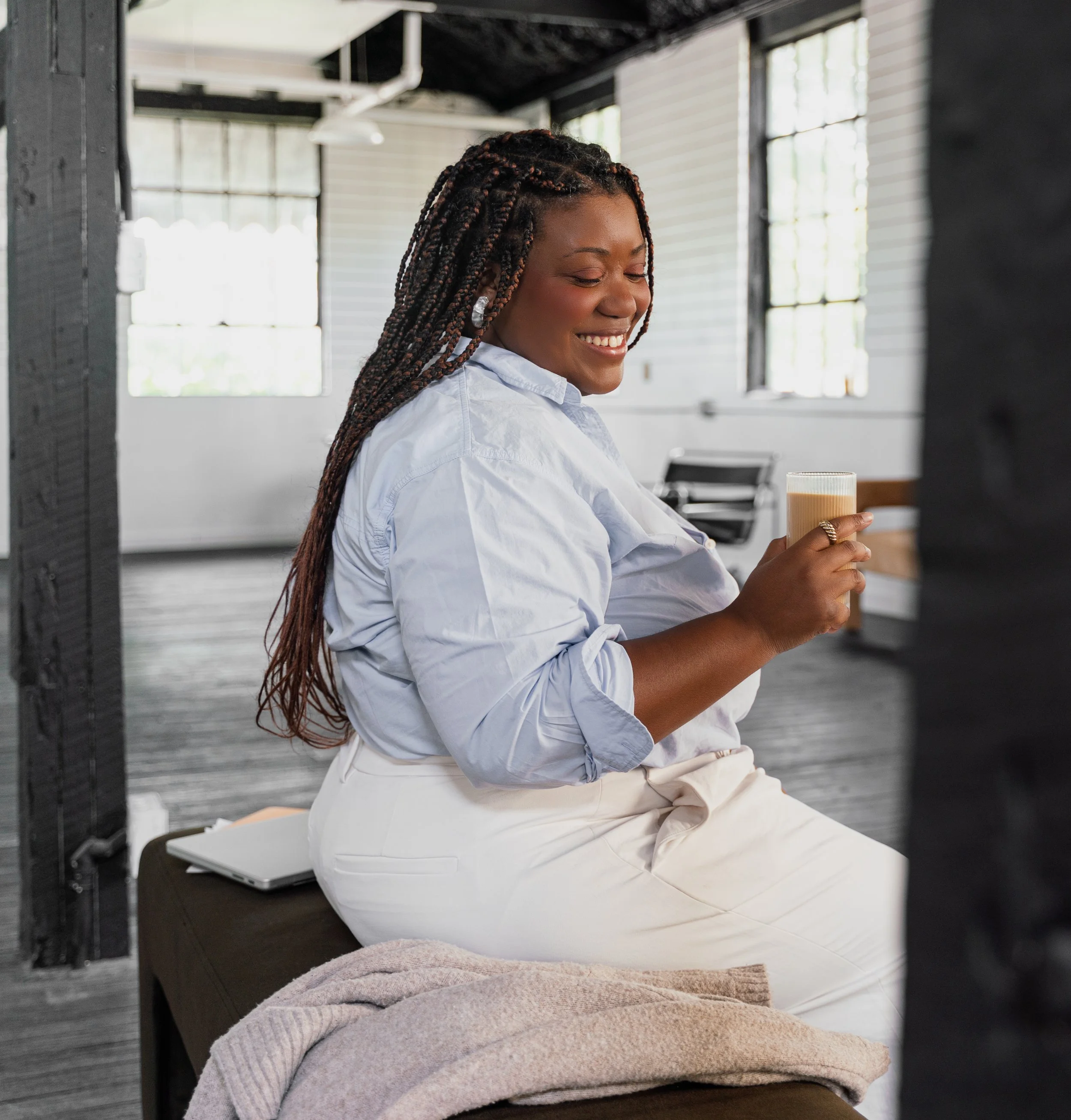 A woman with long braided hair is sitting on a bench, smiling and holding a latte in a bright, modern indoor space with large windows.