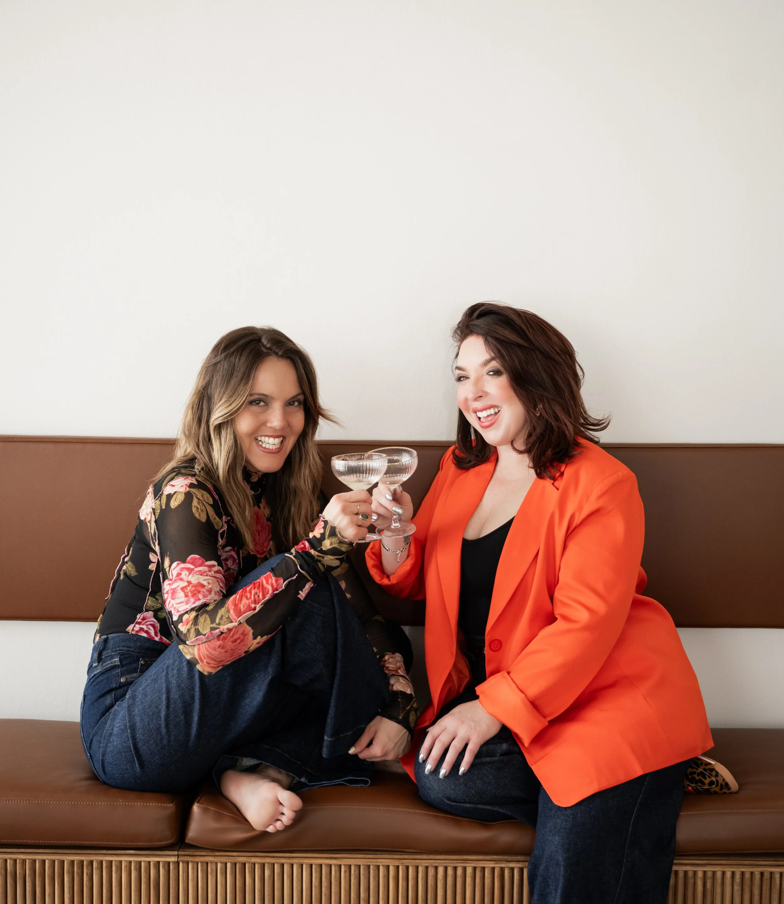 Two women sitting on a brown leather bench, celebrating with cocktails, smiling and toasting.