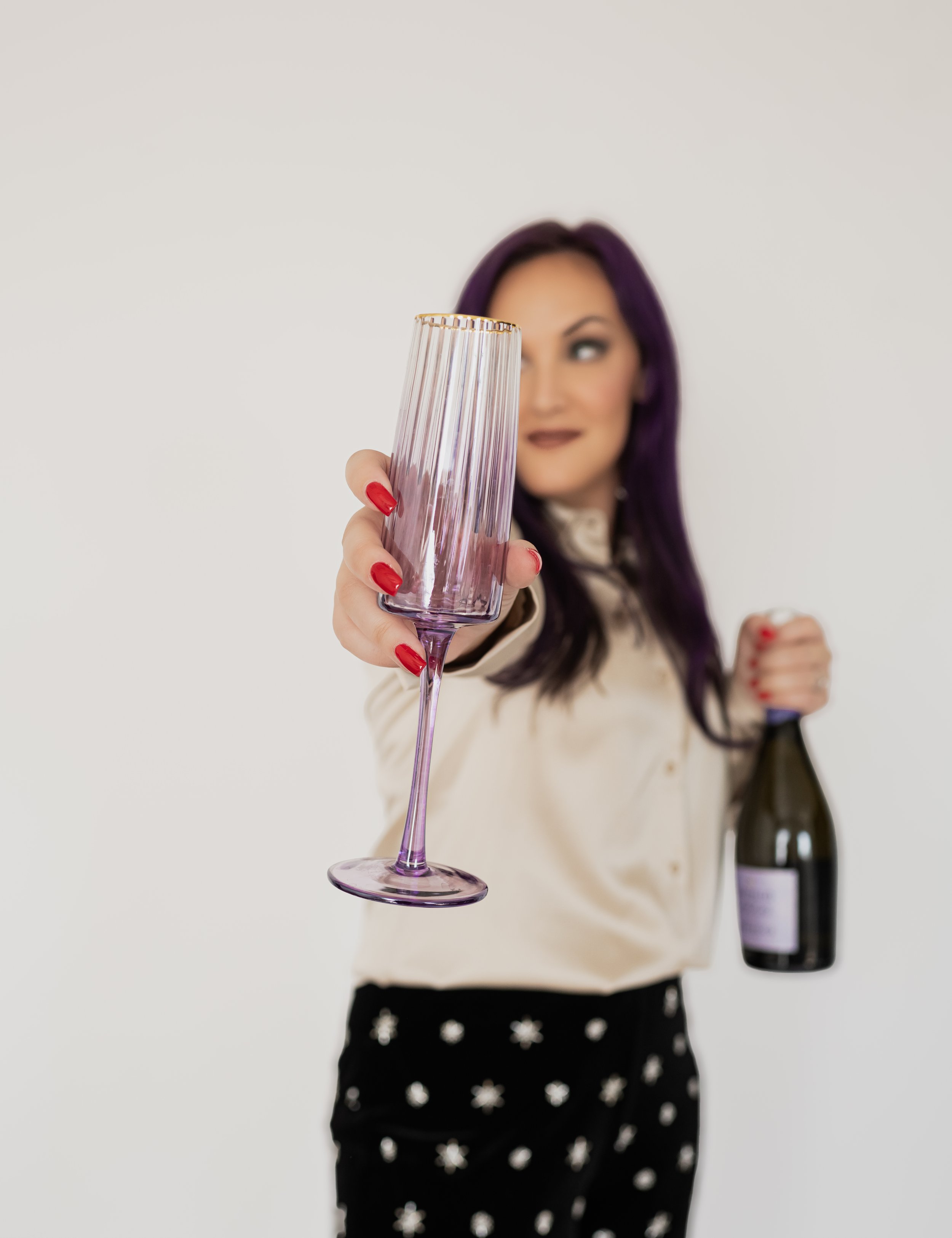 A woman with purple hair holding a purple champagne flute and a bottle of wine, smiling and dressed in a beige silky blouse and black pants with white floral patterns.