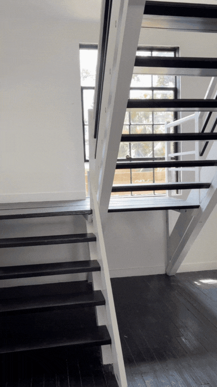 Interior view of a staircase with black steps and white railing, next to large windows with grid pattern, overlooking an outdoor scene with trees.