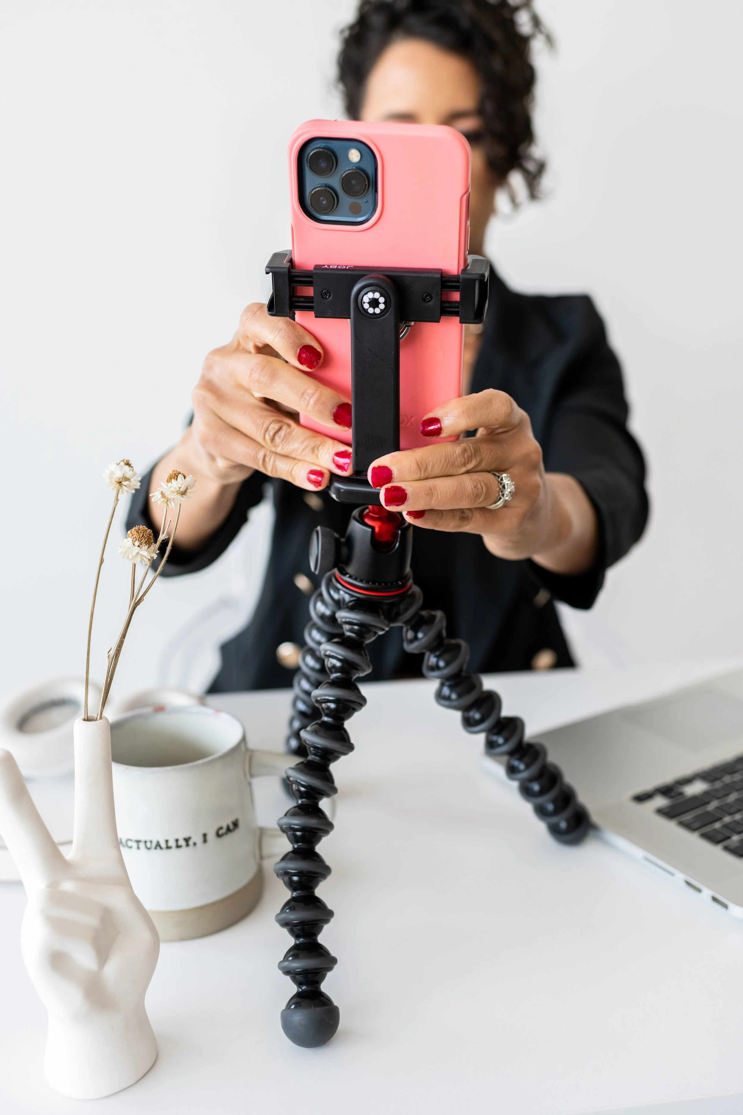 Person with curly hair taking a selfie with a pink smartphone mounted on a flexible tripod on a white desk, with a vase holding dried flowers and a laptop.